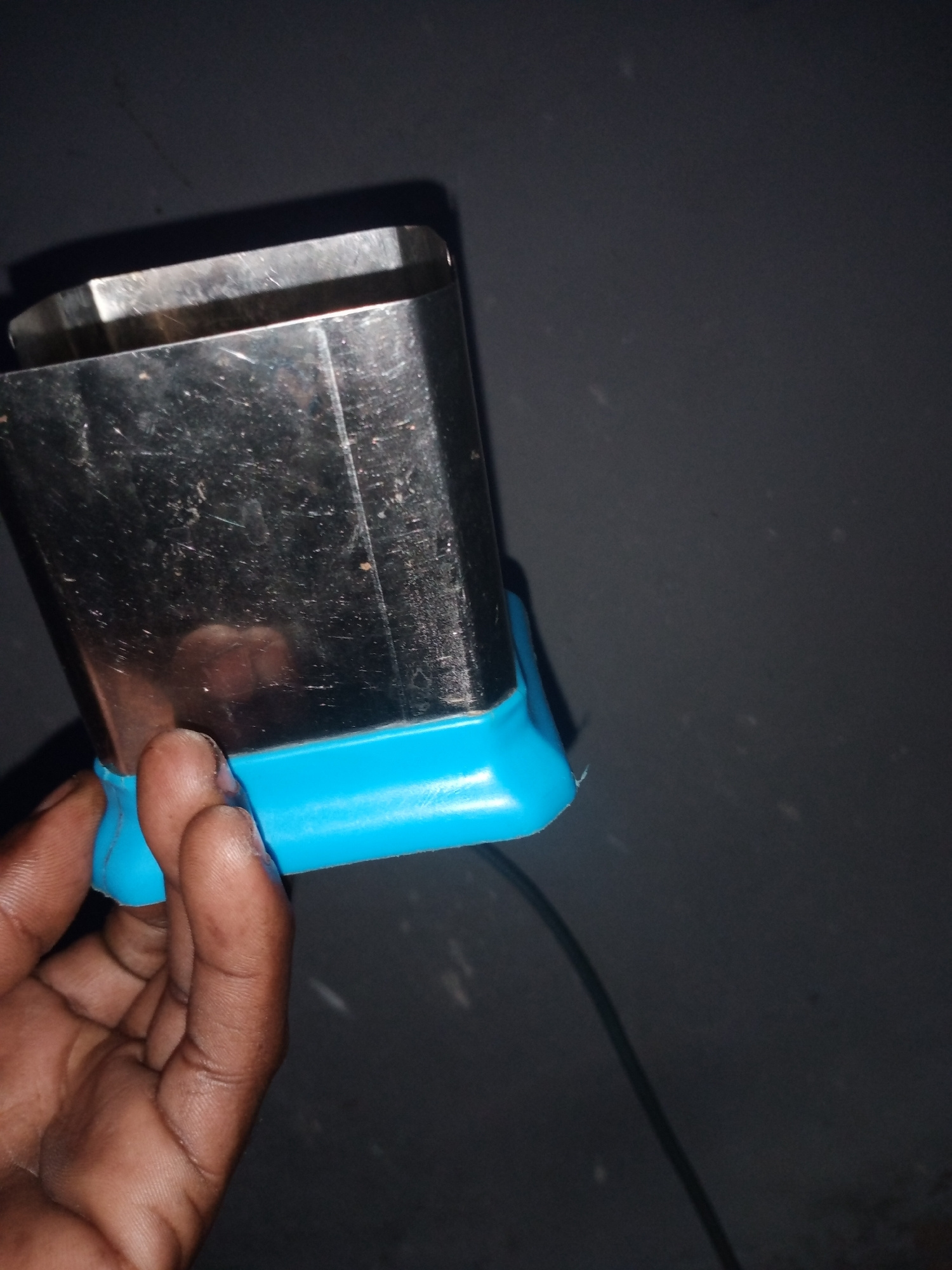 A hand with dark skin is holding a rectangular metal object with a bright blue plastic base. The metal object appears to be a kitchen utensil, possibly a cutter or mold, with visible scratches and smudges on its surface. The background is a dark, textured wall, which could be an exterior wall of a building. The lighting suggests it is indoors or nighttime. This image was taken in Jalingo, Nigeria.