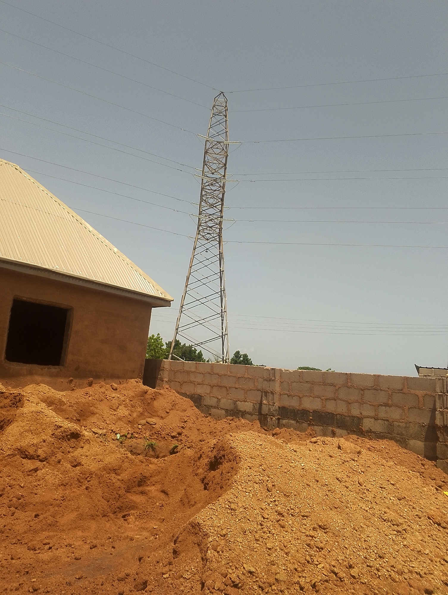 An outdoor, medium shot captures a construction site under a clear, light blue sky. Dominating the right side of the image is a tall, lattice-like transmission tower, its metallic structure stark against the sky. Several parallel power lines stretch horizontally across the frame, originating from the tower and extending towards the left. In the foreground and midground, mounds of reddish-brown soil are piled up, suggesting recent excavation or landscaping. To the left, a partially constructed building is visible, featuring a corrugated metal roof and walls made of a tan, earthy material. One large, dark opening, presumably a window or doorway, is present on the side of the building. A low wall constructed from concrete blocks runs horizontally across the mid-ground, separating areas of the construction site. Lush green trees are visible in the distance behind the block wall, peeking out from behind the unfinished building. The overall scene depicts a developing urban or semi-urban area with active construction activities.
