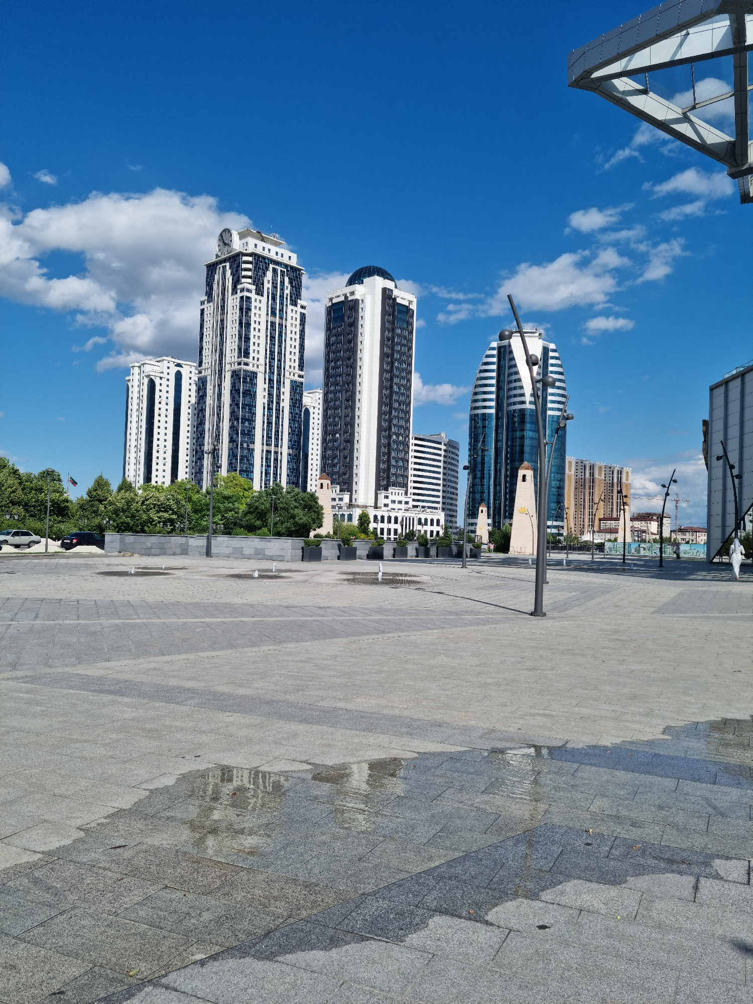 In Grozny, Russia, on a sunny day with scattered clouds, a large, open plaza is depicted. The plaza is paved with large, gray stone tiles, some of which are wet, reflecting the sky and surrounding architecture. In the mid-ground, a line of low-lying fountains sprays water intermittently. Behind the fountains, a stone retaining wall separates the plaza from a green area with lush trees.
Dominating the background are several modern, multi-story buildings. To the left, a tall, white skyscraper with dark vertical accents and a prominent clock face at its peak stands out. Adjacent to it are two other white buildings with similar architectural styles. Further to the right, a distinctive, curved skyscraper with a blue, reflective glass facade and white vertical fins is visible. Another building with a light brown, patterned facade is also present, along with a glimpse of a larger, modern structure on the far right.
Two lampposts are positioned in the plaza, one in the foreground and another further back. The scene suggests a well-maintained urban public space, possibly a square or a park, in a city center. No people are clearly visible, though the presence of a car on the left suggests it is an accessible area. The overall impression is one of contemporary urban development under a bright, blue sky.