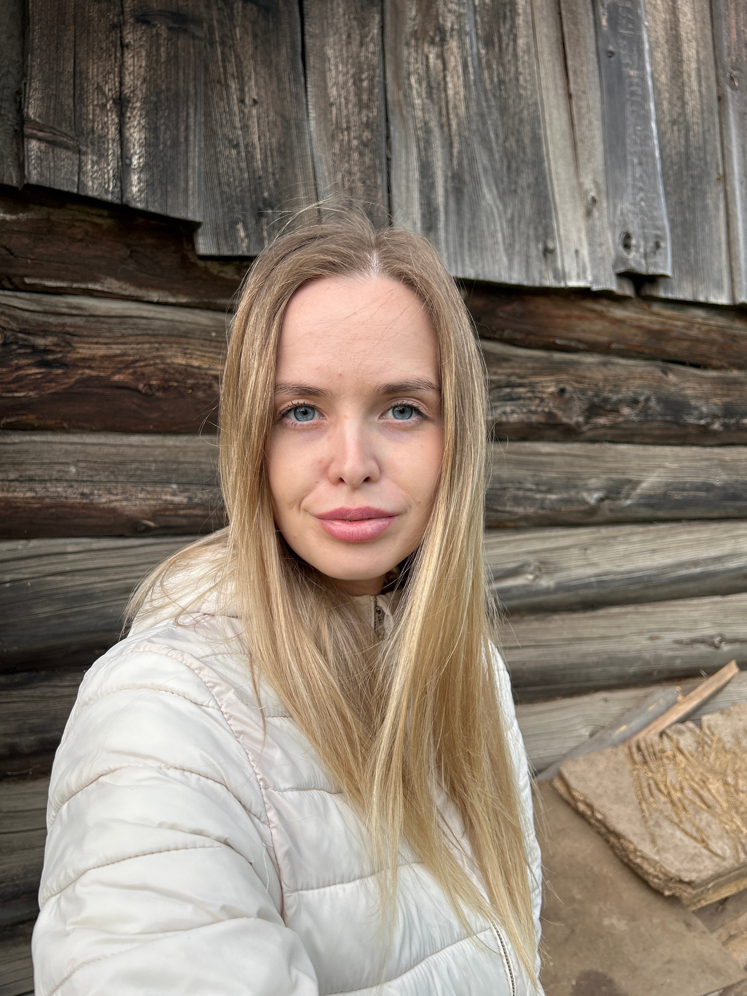 A medium-shot, eye-level, outdoor selfie captures a young Caucasian woman with long blonde hair and blue eyes standing in front of a rustic wooden wall. She is wearing a white quilted jacket. Her gaze is directed at the camera, and her lips are slightly parted, giving her a thoughtful expression. The background is composed of weathered, horizontal wooden planks, some of which appear to be logs. To the right of the woman, a small stack of flat, rectangular objects, possibly wood or cardboard, is visible. The scene appears to be in a rural or historical setting, consistent with a location in the village of Азинский, Russia.