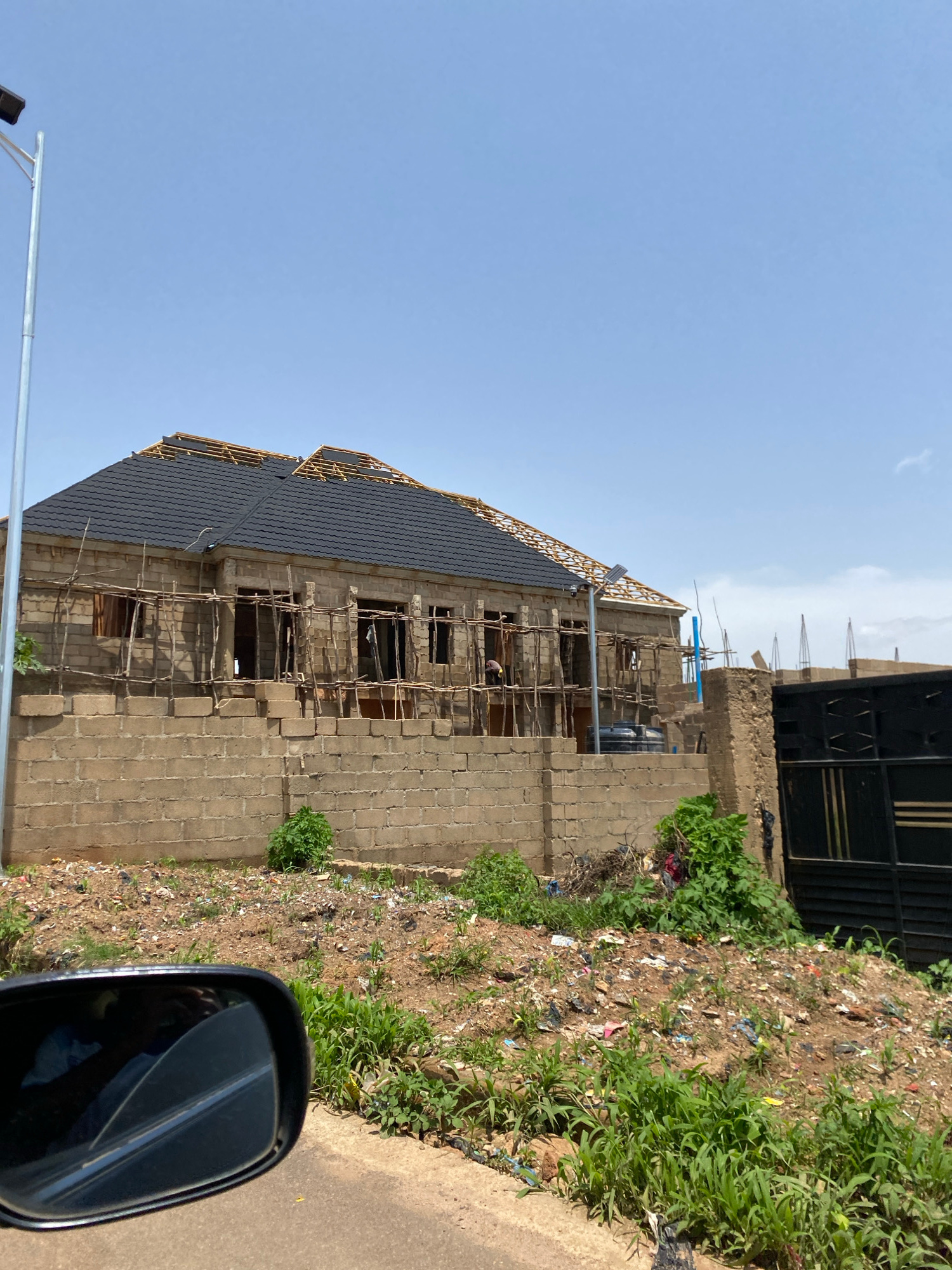 An exterior, eye-level, medium shot captures a one-story building under construction in Gura Top, Nigeria. The building has a newly installed black tiled roof with exposed wooden rafters underneath. The walls are constructed from concrete blocks. Scaffolding made of wood poles is erected around the exterior of the building. Several windows are visible with no glass installed, only wooden supports. A black metal gate is visible to the right of the frame. The foreground is a patch of dry, uneven ground covered in debris, including plastic bags and other refuse. Green grass and weeds are growing sporadically amidst the trash. A portion of a black car side mirror is visible in the lower left corner, reflecting a person inside the vehicle. A light pole with a solar panel is positioned in front of the building. The sky is clear and blue.