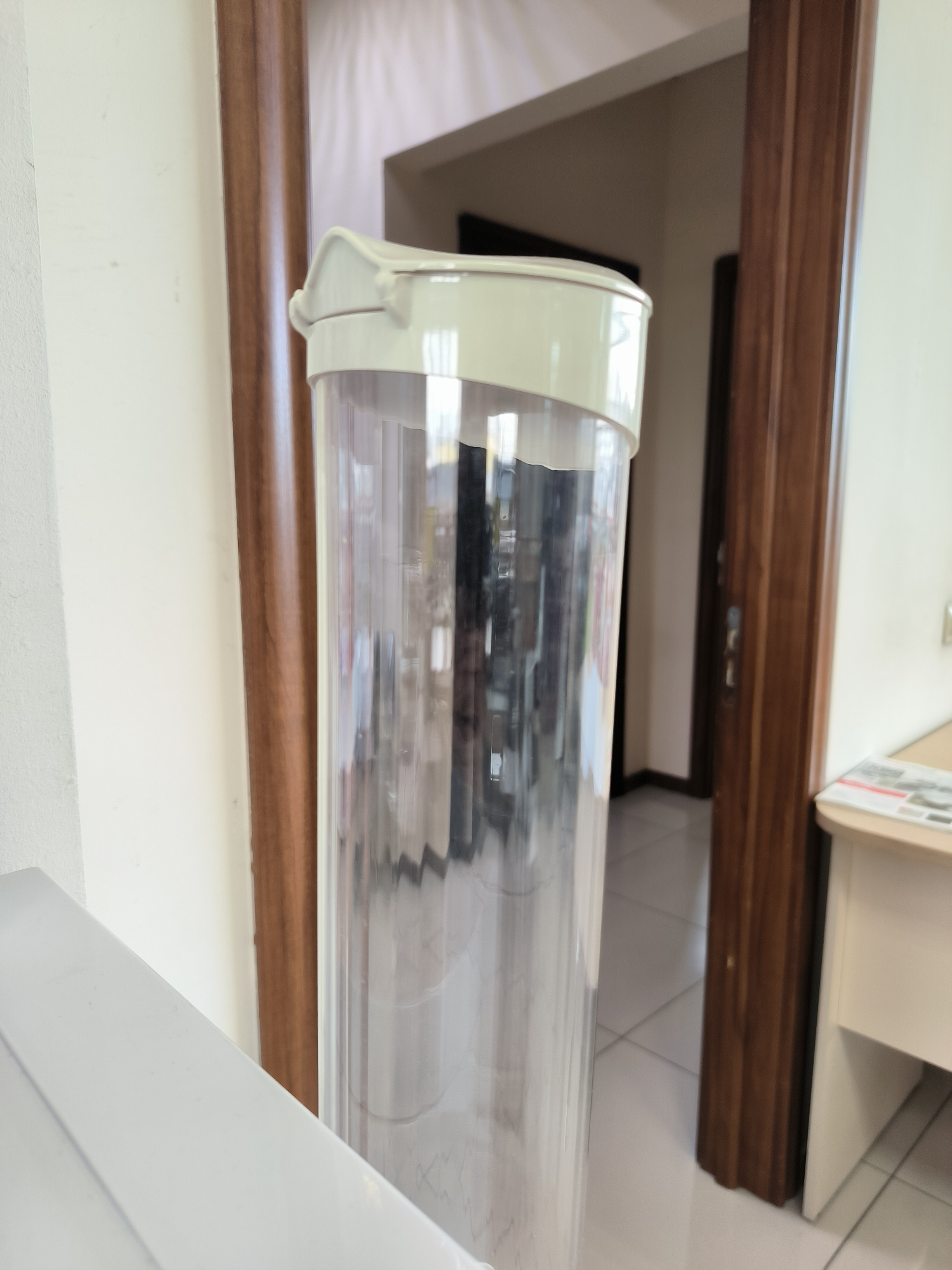 A clear plastic container with a light-colored lid stands vertically in the foreground, its fluted surface visible. The container is positioned to the left of a dark brown wooden door frame, which is partially visible. To the right of the door frame, a portion of a light-colored desk with papers on it is present. The background reveals a room with tiled flooring and another door frame. The scene appears to be indoors, likely in an office or residential setting. The image does not provide specific information about the city of Yuzhno-Sakhalinsk, Russia, or any individuals present.