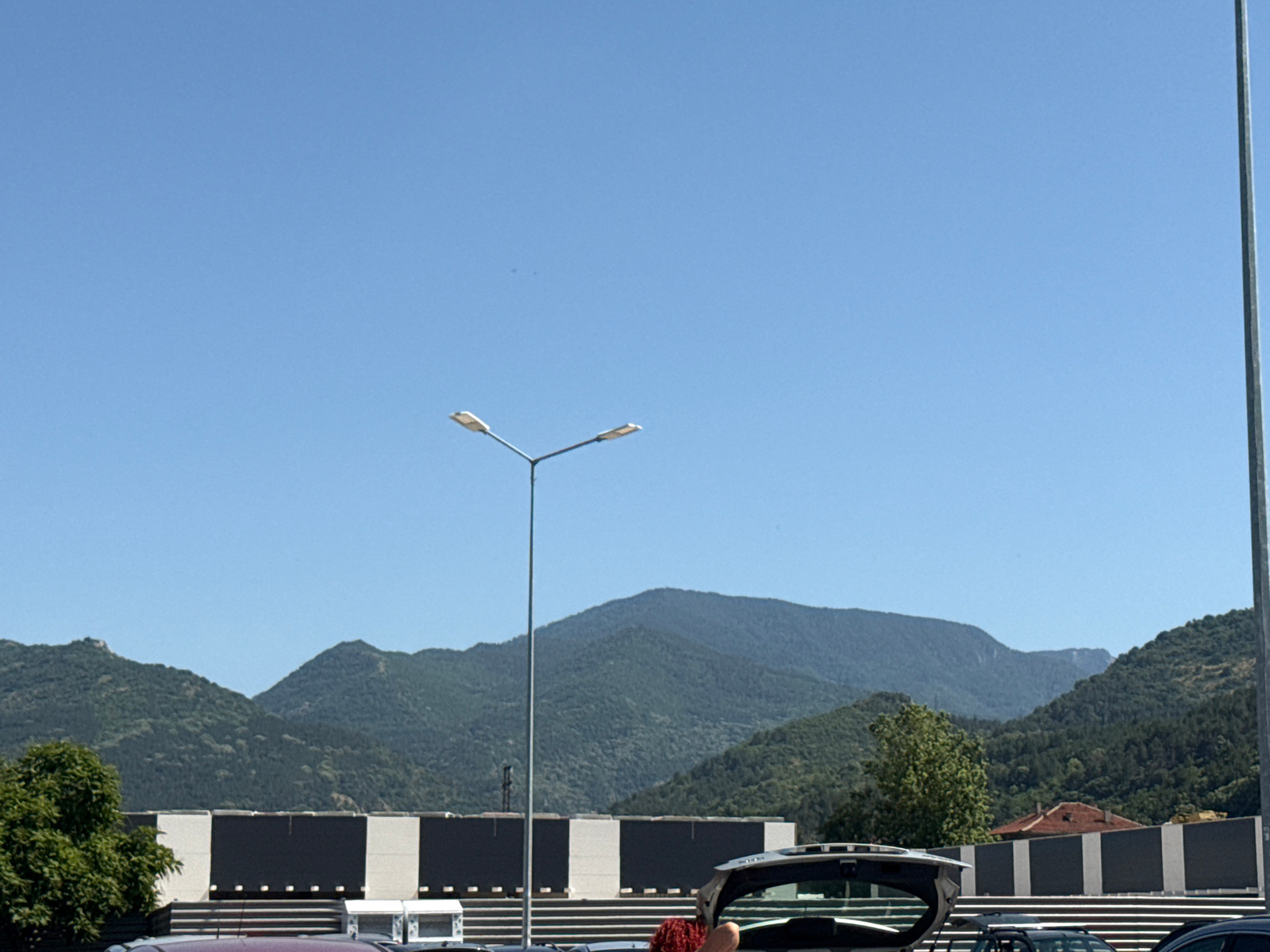 This image depicts a scene in Asenovgrad, Bulgaria, under a clear blue sky. In the foreground, the open trunk of a silver car is visible. Behind the car, a building with a distinct black and white facade is present, likely a commercial or industrial structure, as indicated by the row of evenly spaced doors or loading bays. A street light with two lamps stands prominently in the middle ground, its metallic pole extending upwards. Beyond the building, a range of lush green mountains forms the backdrop, their slopes covered in dense foliage. The overall environment suggests an outdoor setting, possibly a parking lot or an area adjacent to a business establishment, with the mountains providing a natural scenic element.