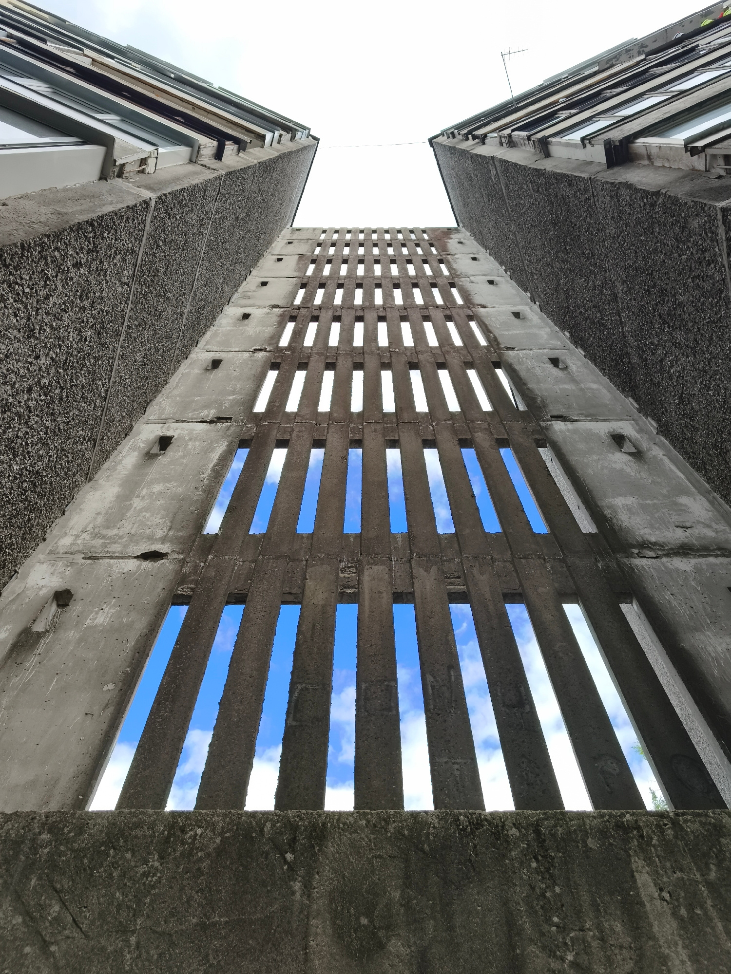 The image is a low-angle shot looking up between two brutalist concrete buildings. The buildings' textured facades create a stark, geometric contrast. A central concrete structure, also textured, is pierced by numerous narrow, vertical openings that reveal a bright blue sky with scattered white clouds. These openings are arranged in a rhythmic pattern, forming a sort of slatted facade. The overall impression is one of architectural austerity and a sense of looking upwards towards the heavens. The time of day appears to be daytime, given the bright sky. The weather seems pleasant, with a clear blue sky and clouds. There are no people or specific objects visible beyond the architecture itself. No text is discernible in the image. The location is unknown.