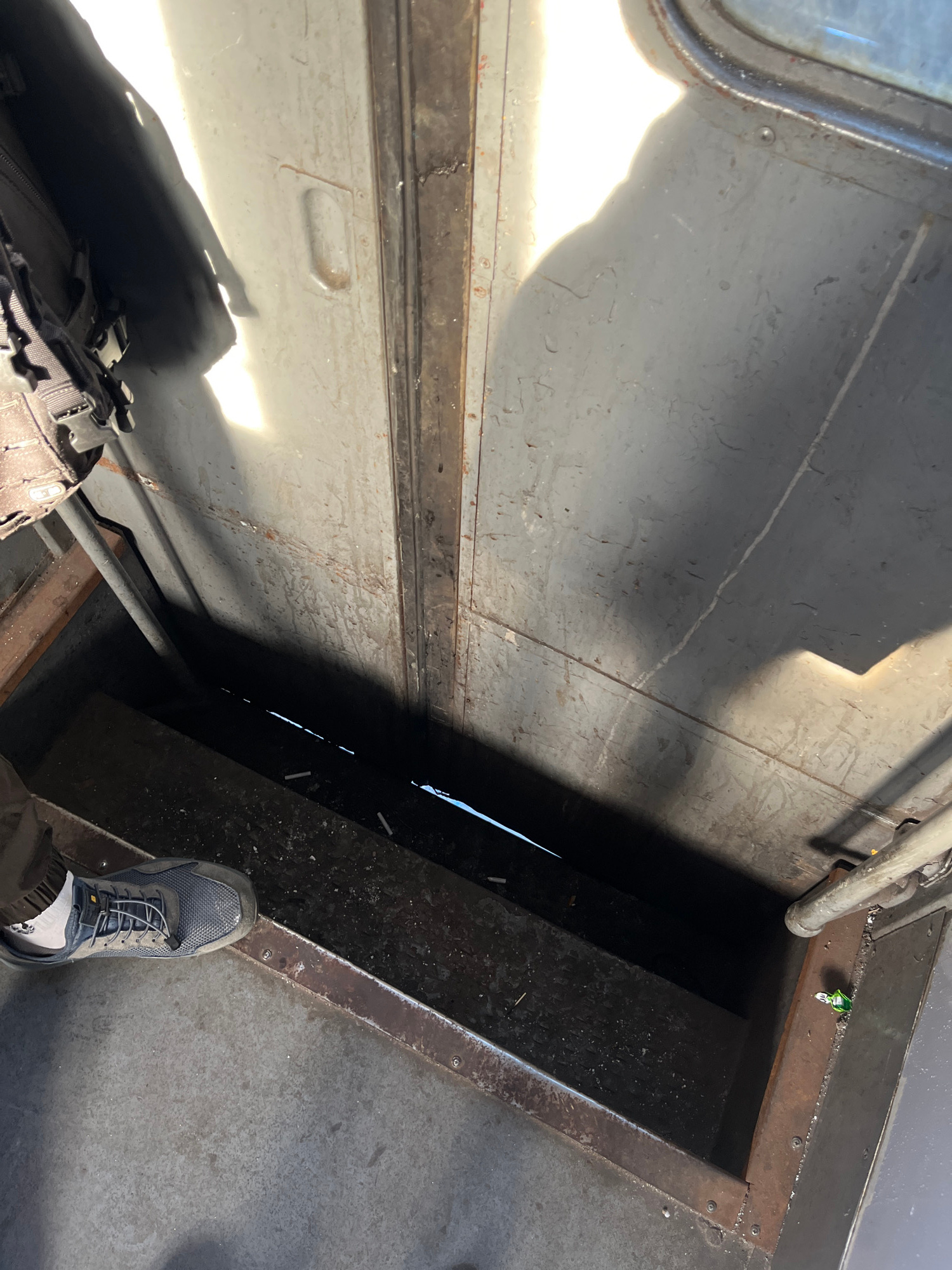 This is an eye-level, close-up shot taken from above, looking down into a dark compartment. The scene is illuminated by a strong light source casting shadows, suggesting it is daytime and outdoors or near a window. In the foreground, a person's left foot wearing a dark grey athletic shoe with grey laces is visible, standing on a textured metal floor. The shoe is positioned at an angle, pointing towards the lower left of the frame. The floor appears to be a grey, metallic surface with some textured markings.
The compartment below is dark and rectangular, with what appear to be steps or ledges leading downwards. A metal pipe runs along the top edge of the compartment. In the dark area, several small, white cylindrical objects are scattered, potentially cigarette butts. The walls of the compartment are dark and metallic, with visible seams and some rust.
To the left and extending out of the frame, a portion of a black backpack with buckles and straps is visible, hanging off the edge. The background is dominated by a large, grey, metallic surface, possibly a door or wall, with a recessed handle or latch. The lighting creates strong contrasts, with bright areas of light and deep shadows. The overall environment appears industrial or utilitarian, possibly part of a vehicle, building, or outdoor structure. The image does not provide any specific clues to its location within the city of Kyiv, Ukraine.