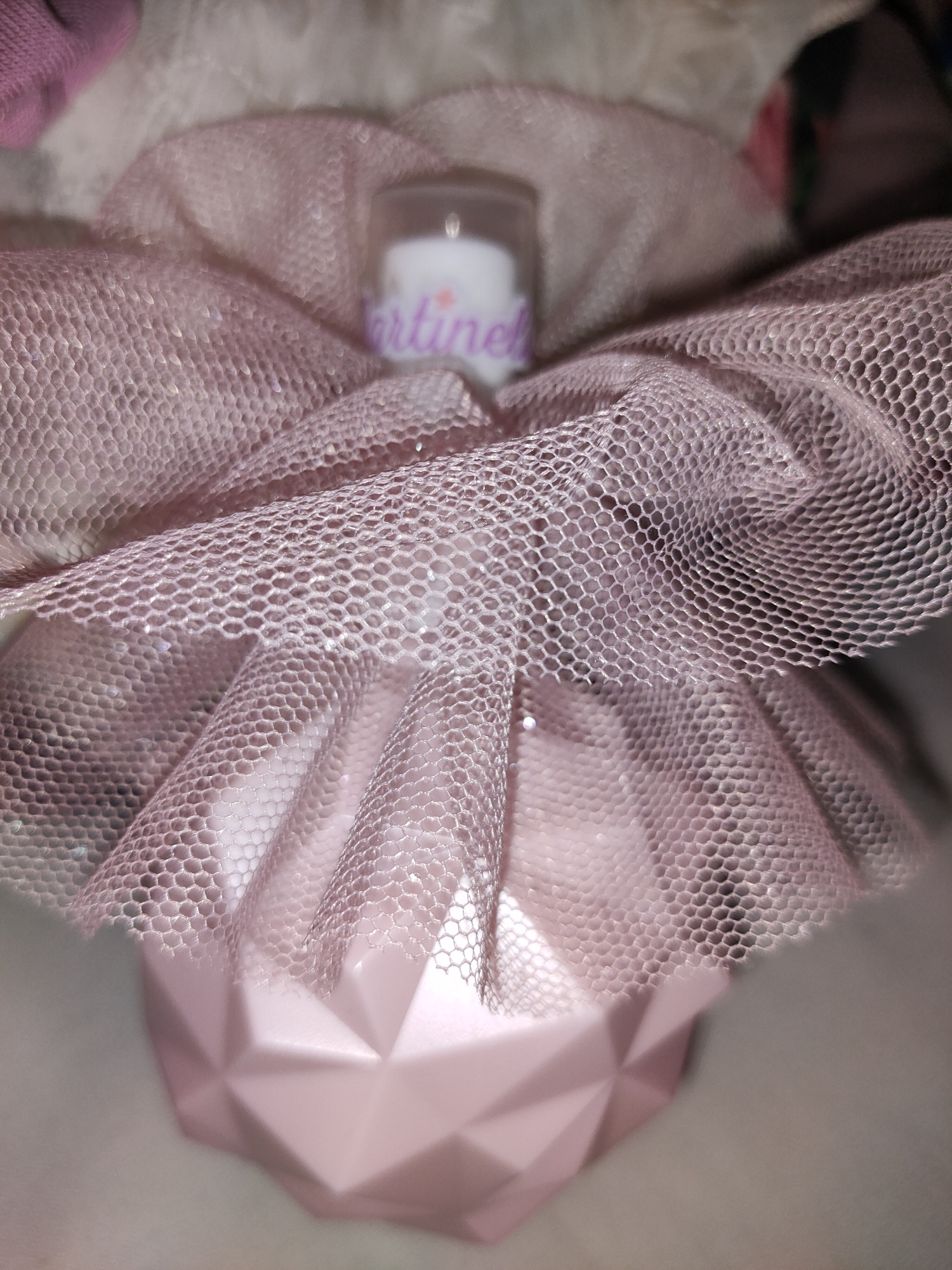 The image displays a close-up, slightly blurred view of a pink, faceted object, possibly a perfume bottle, adorned with a delicate, light pink, hexagonal-patterned netting that creates a tutu-like effect around the top. A small, clear cylindrical object with a white label, partially obscured by the netting and text that appears to read "martine", is visible at the center of the netting. The background is soft and out of focus, with hints of pale fabric and a darker, indistinct element on the right. No specific location in Perpignan, France, can be identified from this image.