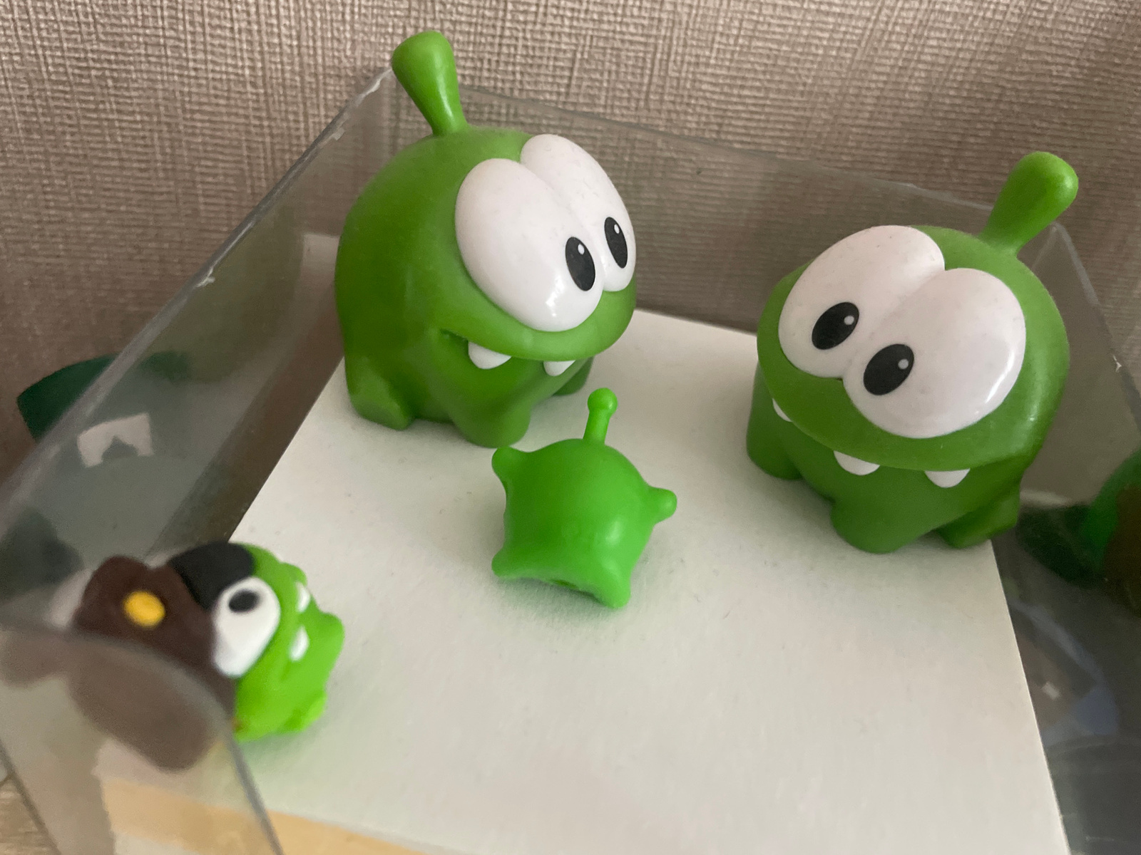 This media file features a close-up, eye-level shot of several small, green, cartoon-like figures with large white eyes and black pupils. There are three main figures prominently displayed, resembling characters from the game "Cut the Rope." Two larger figures are positioned on the right and left sides of the frame, facing inwards. A smaller, almost star-shaped figure sits in the center, between the two larger ones. A fourth, partially visible figure is in the lower left corner, appearing to be a brown and green character with a yellow dot. The figures are placed on a white surface, possibly paper or a placemat, which is enclosed within a clear plastic container. The background consists of a textured, light brown material, resembling a woven fabric or wallpaper. The lighting appears to be artificial, suggesting an indoor setting. The overall scene is a still life arrangement of these collectible figures, with no discernible activity or event taking place. The mood is playful and lighthearted, evoked by the cartoonish design of the subjects. There is no visible text in the image. The location context is Moscow, Russia.