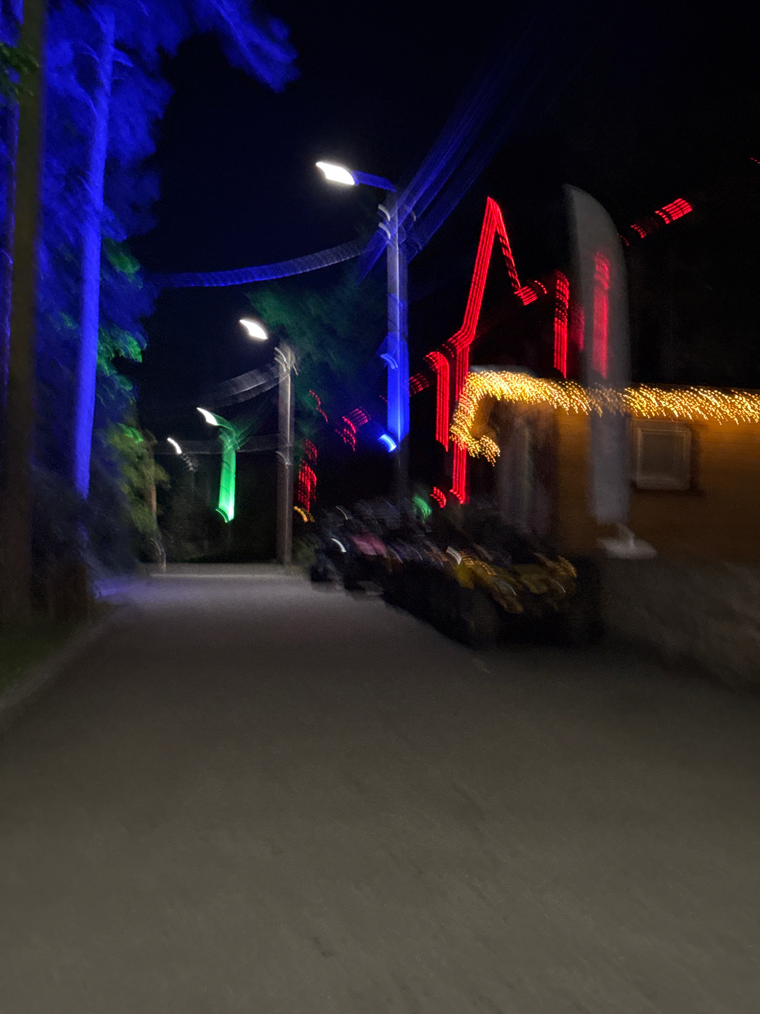 The image depicts a blurred nighttime scene with various lights and structures. On the left, trees are illuminated with bright blue light. Further down the path, there are streetlights, some of which emit white light, and one appears to have green light. On the right side of the image, a building is visible, decorated with a string of warm yellow lights along its roofline. A red, angular light structure is also present near the building. In the foreground, a dark, blurry object that could be a vehicle or a group of people is present on the path. The scene suggests an outdoor public space, possibly a park or a street decorated for an event. The overall impression is one of movement and vibrant, artificial illumination against a dark background.