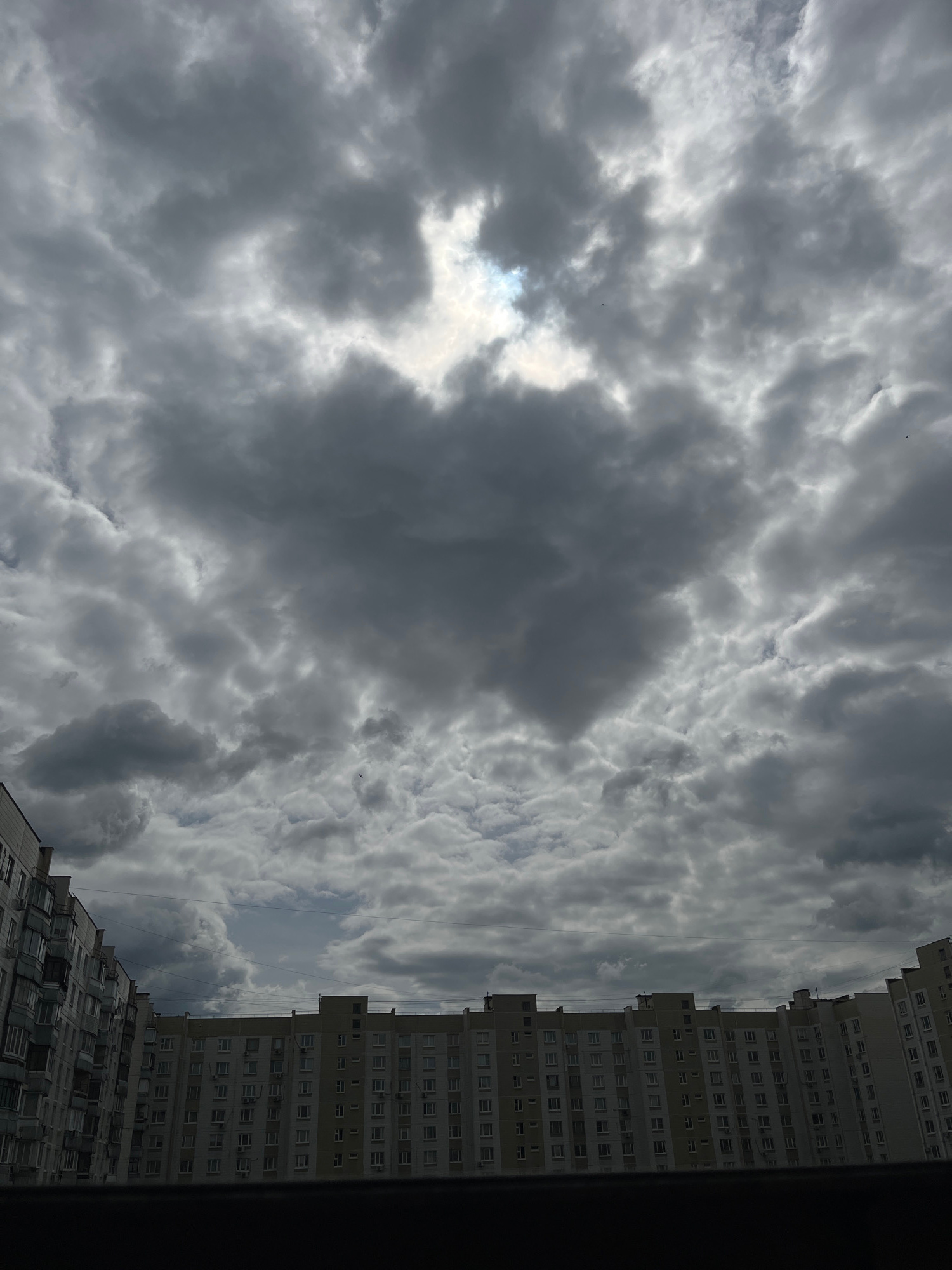 The image displays a dramatic sky filled with dense, dark grey clouds, characteristic of an impending storm or overcast weather. A small patch of bright light pierces through the clouds near the center-top of the frame, suggesting the sun is attempting to break through. In the lower portion of the image, a series of identical, multi-story apartment buildings are visible. These buildings have a light-colored facade with rows of windows and balconies, and they appear to be constructed in a uniform style, suggesting they are part of a large housing development. The perspective suggests the photo was taken from a slightly elevated position, looking up towards the sky and across the rooftops of the buildings. The overall scene is urban, with a focus on the contrast between the imposing natural elements of the sky and the man-made structures of the residential buildings. There are no people or active events depicted in the image, creating a still, atmospheric scene. The specific location within the city of Moscow, Russia, cannot be determined from the visual information alone, as the architecture is generic to many Soviet-era housing blocks found throughout Russia and Eastern Europe.