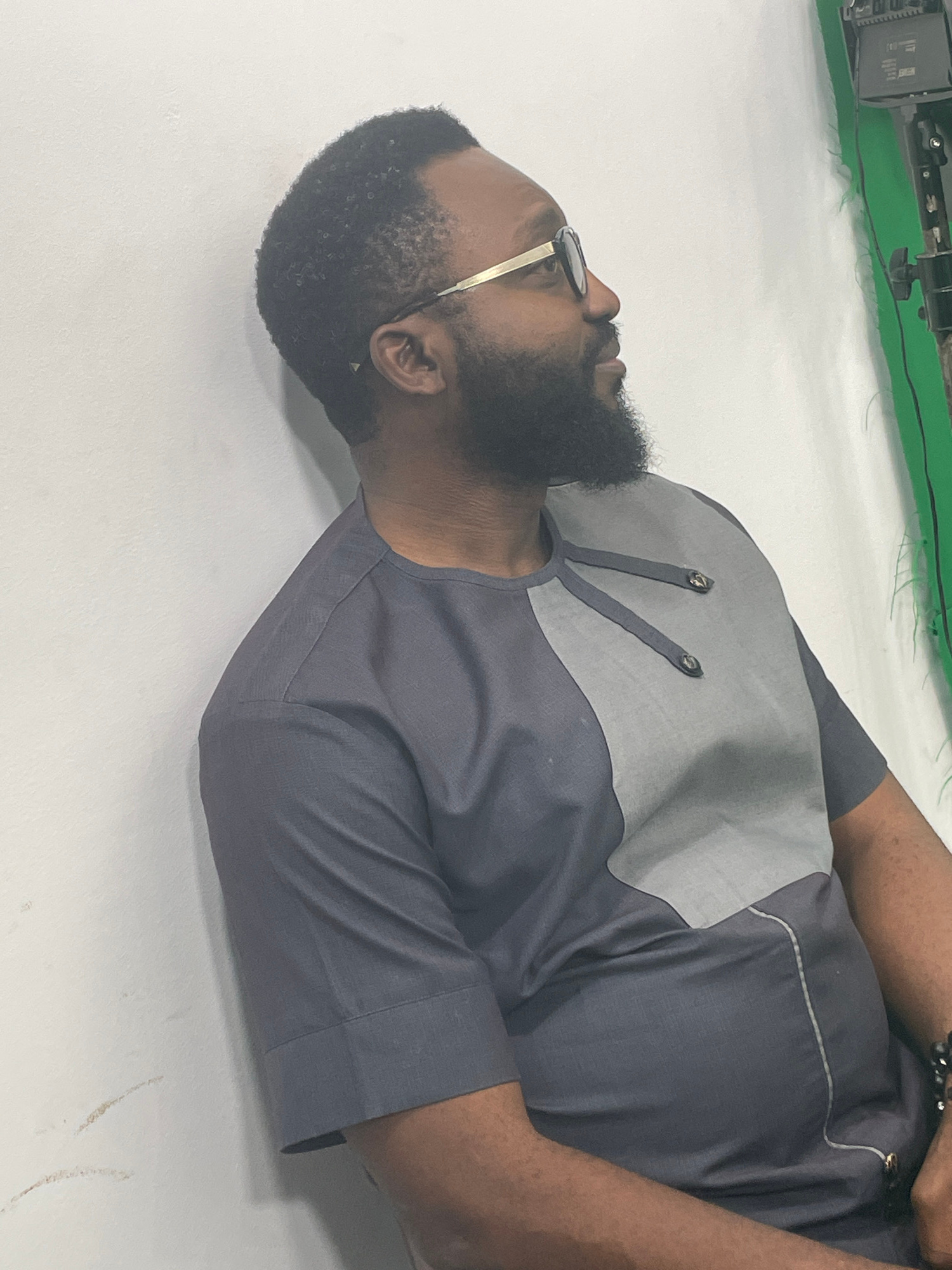 The image shows a man lying down, facing left, with his head slightly tilted up. He has dark skin, a beard, and is wearing sunglasses with gold frames. He is dressed in a grey two-toned shirt with a lighter grey panel on the chest, featuring decorative stitching and two metallic buttons. His arms are resting by his sides. The background appears to be a white surface with some faint markings, and a green backdrop with some equipment is visible at the top left. The lighting suggests an indoor setting. The orientation of the image is portrait, with the man positioned on the right side of the frame.