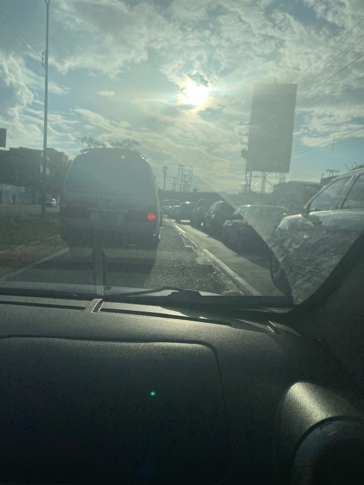The image shows a traffic jam viewed from inside a car, looking through the windshield. The sky is bright with the sun shining through clouds, creating a lens flare effect. Several vehicles are visible on the road, including a dark colored van in the foreground, a silver SUV to the right, and other cars in the distance. There are street lights and overhead power lines visible. In the background, there are buildings and a large billboard structure. The scene appears to be on a highway or major road within the city of Maracay, Venezuela, likely during daylight hours. The overall atmosphere is one of urban congestion under a bright, partially cloudy sky.