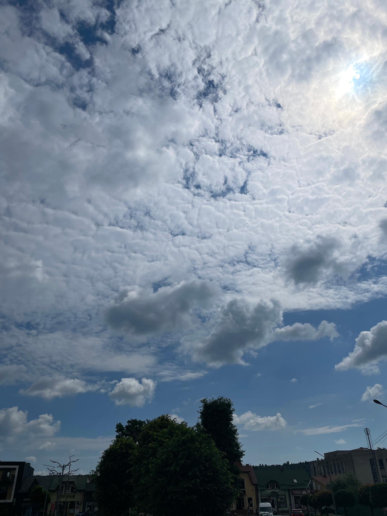 The image shows a sky filled with cumulus clouds and some cirrocumulus clouds. The sun is visible in the upper right corner, partially obscured by clouds. Below the sky, there are trees and buildings, suggesting an urban or suburban environment. There are no people visible in the image. The scene appears to be outdoors during the day.