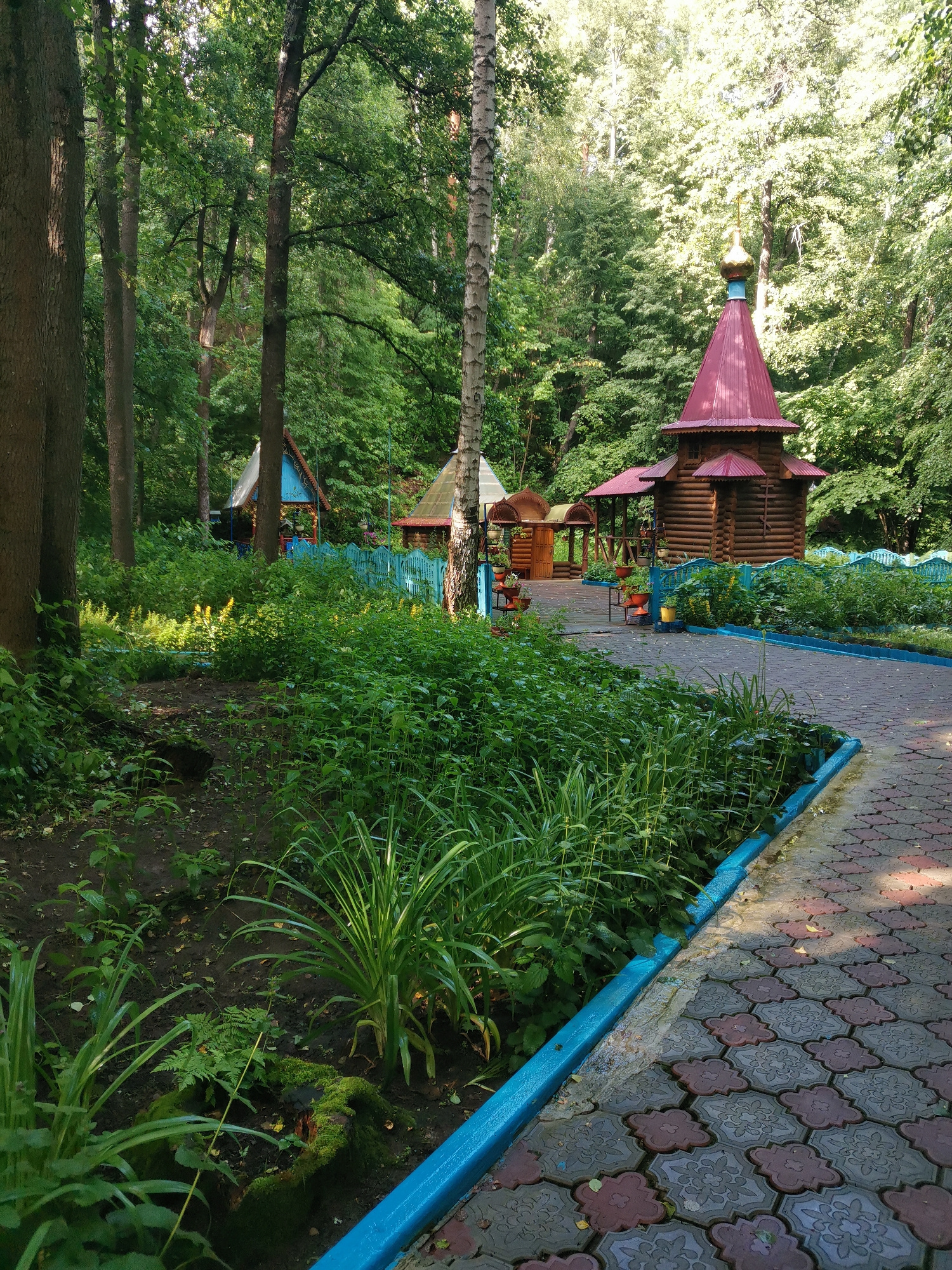 The image shows a paved pathway in a park-like setting. The pathway is made of interlocking red and grey pavers and is bordered by a bright blue curb. To the left of the pathway, there is a dense growth of green foliage, including ferns and tall grass. In the background, there are several wooden structures, including a small, red-roofed chapel with a golden dome, and other small buildings with blue and red roofs. The area is surrounded by tall trees with lush green leaves, suggesting a natural or wooded environment within a city. The lighting indicates it is daytime, possibly morning or afternoon, with sunlight filtering through the trees. There are no people or animals visible in the image.