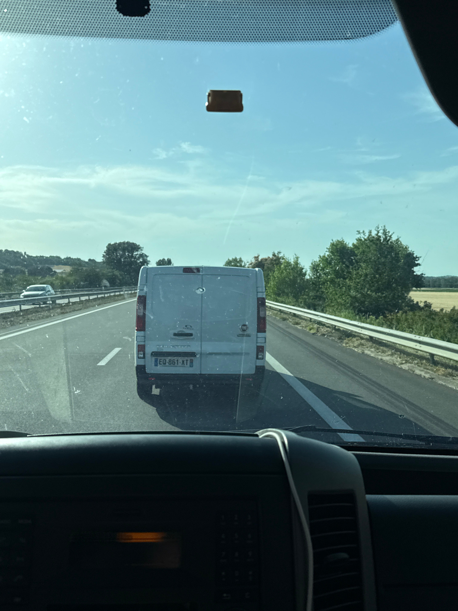 The image is a forward-facing view from inside a vehicle, looking out through the windshield. The vehicle appears to be on a highway. In the foreground, the dashboard of the vehicle is visible, with a dark, textured surface and a faint orange glow from an indicator light. The windshield has some dust or smudges on it.
Outside the vehicle, the highway stretches ahead, with a white line marking the lane. To the left, a silver car is visible on the highway, in a lane further away. A guardrail separates the highway from the grassy shoulder and vegetation.
In the middle ground, a white panel van is directly ahead, traveling in the same direction. The van has a license plate that reads "EQ-861-XT". To the right of the van, there is a dense line of trees and foliage. Beyond the trees, a field of dry, golden crops is visible under a bright, partly cloudy sky. The overall impression is a sunny day in a rural or semi-rural setting. The location is specified as Mas-Saintes-Puelles, France.
