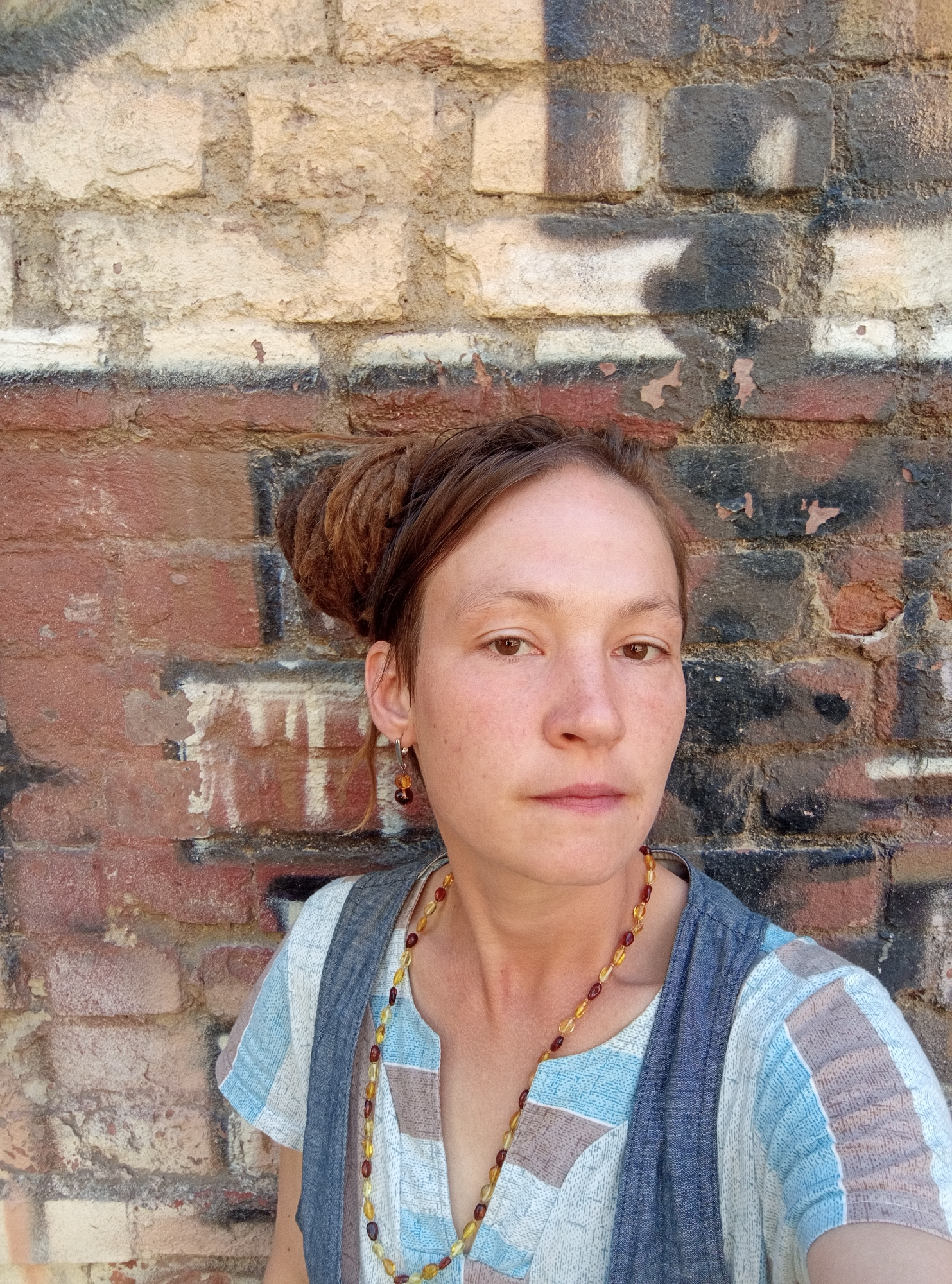 A medium shot shows a woman with dreadlocks pulled up in a bun, facing the camera directly. She has fair skin with freckles, brown eyes, and light brown lips. She is wearing a striped blue and white shirt and a grey vest, with a necklace of amber-colored beads. She has a single dark bead earring on her left ear. The background is a brick wall with graffiti, showing signs of wear and tear. The overall lighting suggests an outdoor setting on a sunny day. The specific location in Saint Petersburg, Russia is not determinable from the provided image.