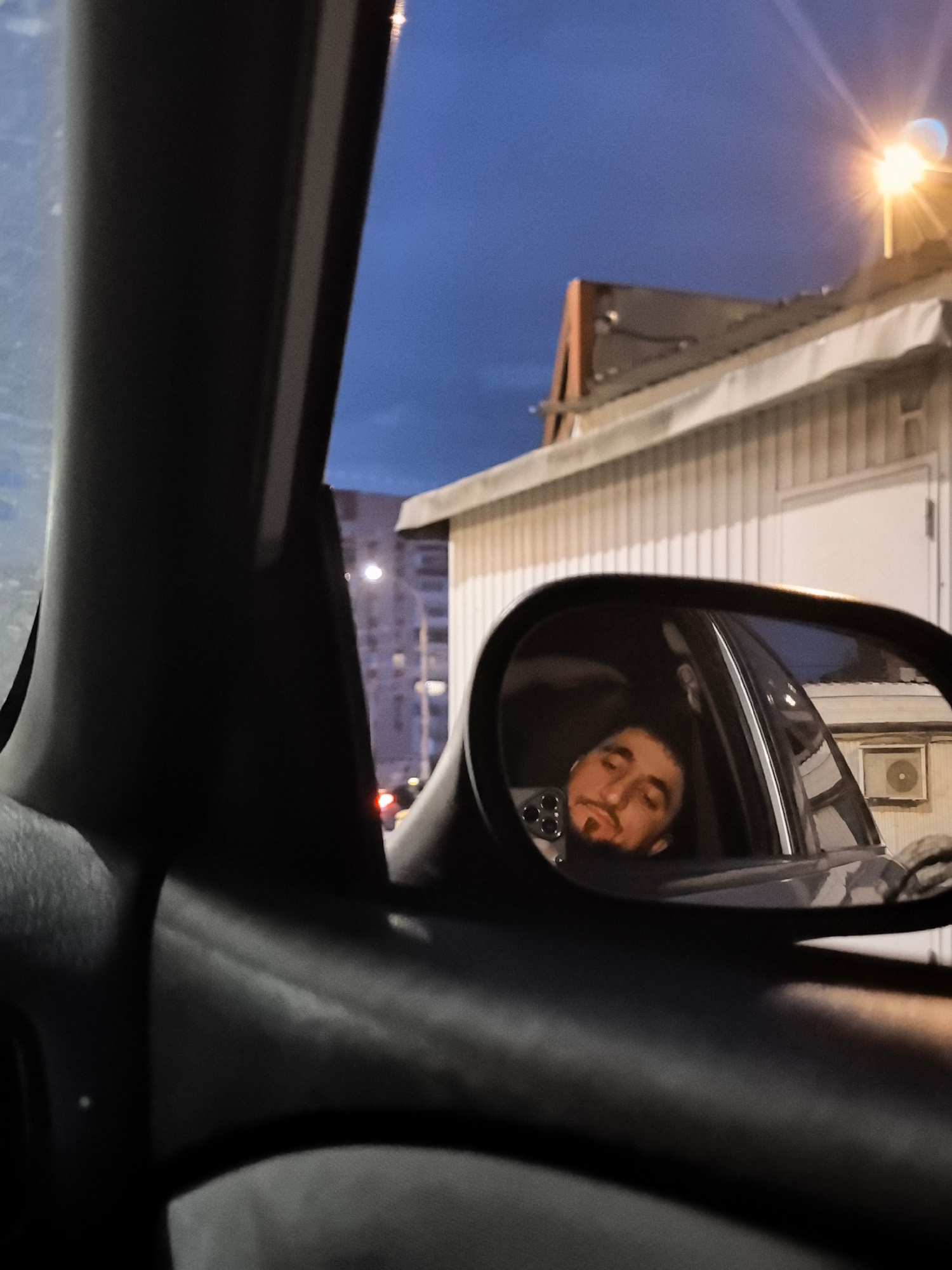 The image is a close-up view from inside a car, looking out through the side window and the side-view mirror. The side-view mirror reflects the face of a man who appears to be sleeping or resting, with his eyes closed and head tilted. He has a beard and is wearing earbuds. The interior of the car is dark. Outside the car, the scene depicts a twilight sky with a bright street lamp illuminating a building with corrugated siding. In the background, a multi-story residential building is visible. The scene appears to be in an urban environment, possibly a parking lot or street in the city of Ivanovo, Russia. The overall atmosphere is calm and still, suggesting a moment of rest.