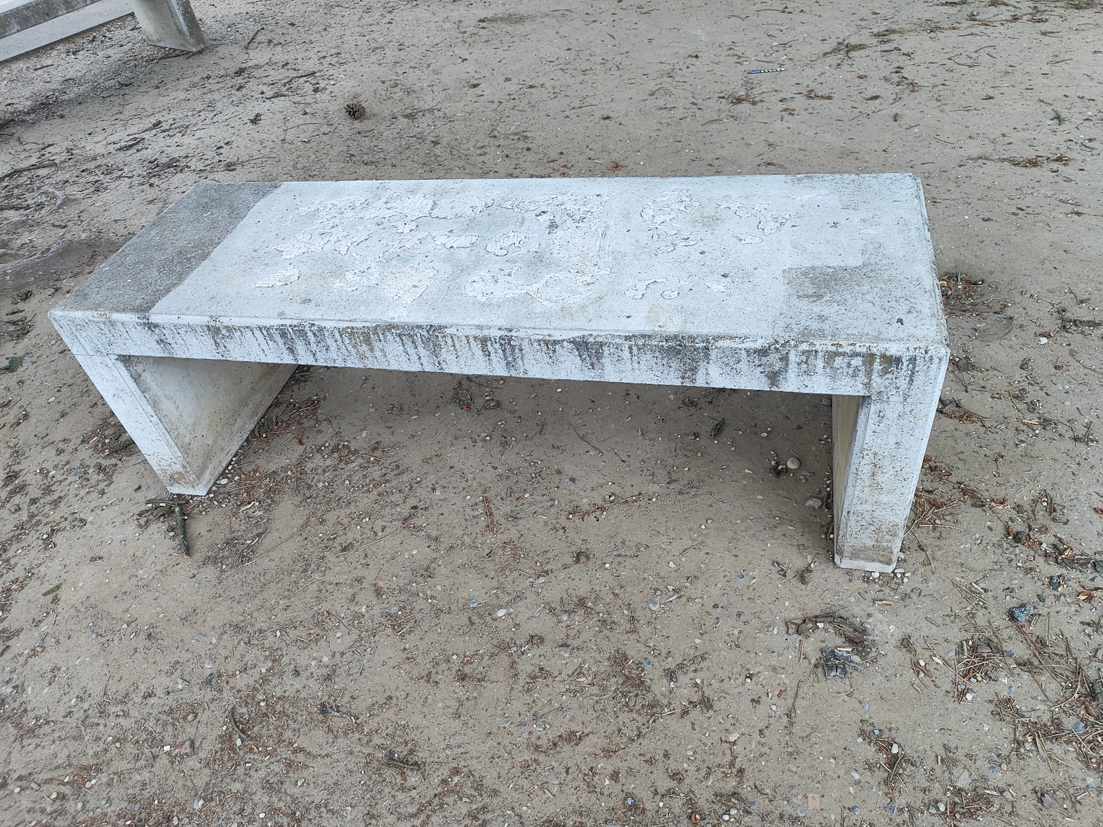 A weathered, concrete bench sits on a sandy, slightly gritty ground surface scattered with small debris such as twigs and pine needles. The bench has a rough, unpainted texture, with areas of peeling paint and staining. The ground appears to be an outdoor public space, possibly a park or recreational area in Diedorf, Germany. The lighting suggests it is daytime. No people or animals are visible in the frame.