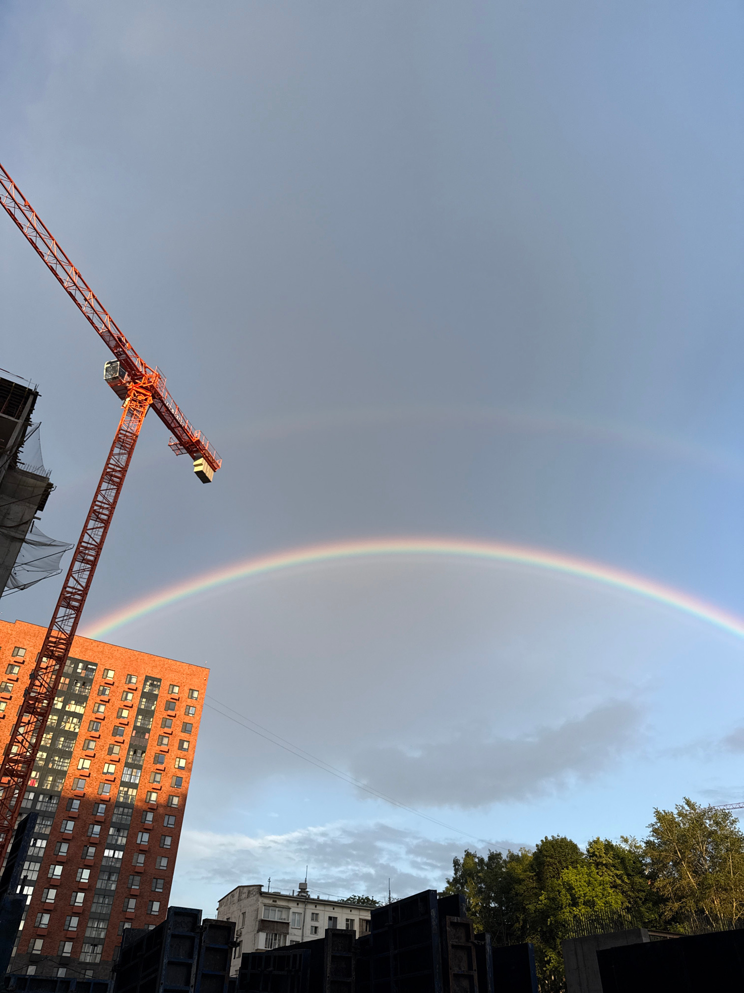An image captures a construction site with a vibrant double rainbow arching across a cloudy sky in Moscow, Russia. In the foreground, a tall, orange construction crane is positioned on the left side of the frame. To its right, a modern, multi-story brick apartment building with numerous windows stands prominently, catching the sunlight on its facade. In the background, other buildings and lush green trees are visible. The sky is a mix of light blue and gray clouds, with the sun casting a warm glow on the scene. Dark, heavy construction materials are also visible in the lower portion of the image.