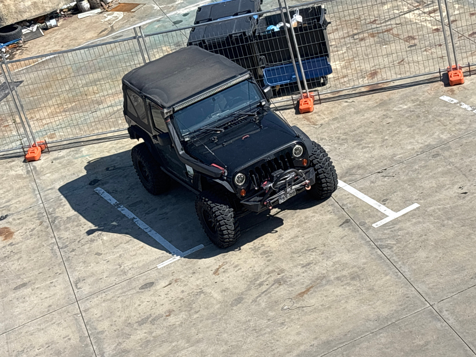 An elevated, high-angle medium shot captures a black Jeep Wrangler parked on a concrete lot with white parking lines. The Jeep is positioned diagonally in the frame, with its front facing slightly left. It has a black soft top, a light bar mounted above the windshield, and large, knobby tires. A license plate reading "288 RR" is visible on the front.
Behind the Jeep, a temporary chain-link fence separates the parking area from a construction or industrial zone. Orange plastic barriers are visible at the base of the fence posts. In the background, behind the fence, are two large black crates or containers, one with a blue lid, and some discarded items including a tire. The ground surface appears to be worn concrete with some water stains and dirt marks. The scene is lit by natural sunlight, casting shadows from the Jeep and the fence onto the concrete. No people are visible in the image. The context suggests an outdoor area, possibly a construction site or a storage yard, which could be located in the city of Tbilisi, Georgia, given the specific location mentioned in the prompt.
