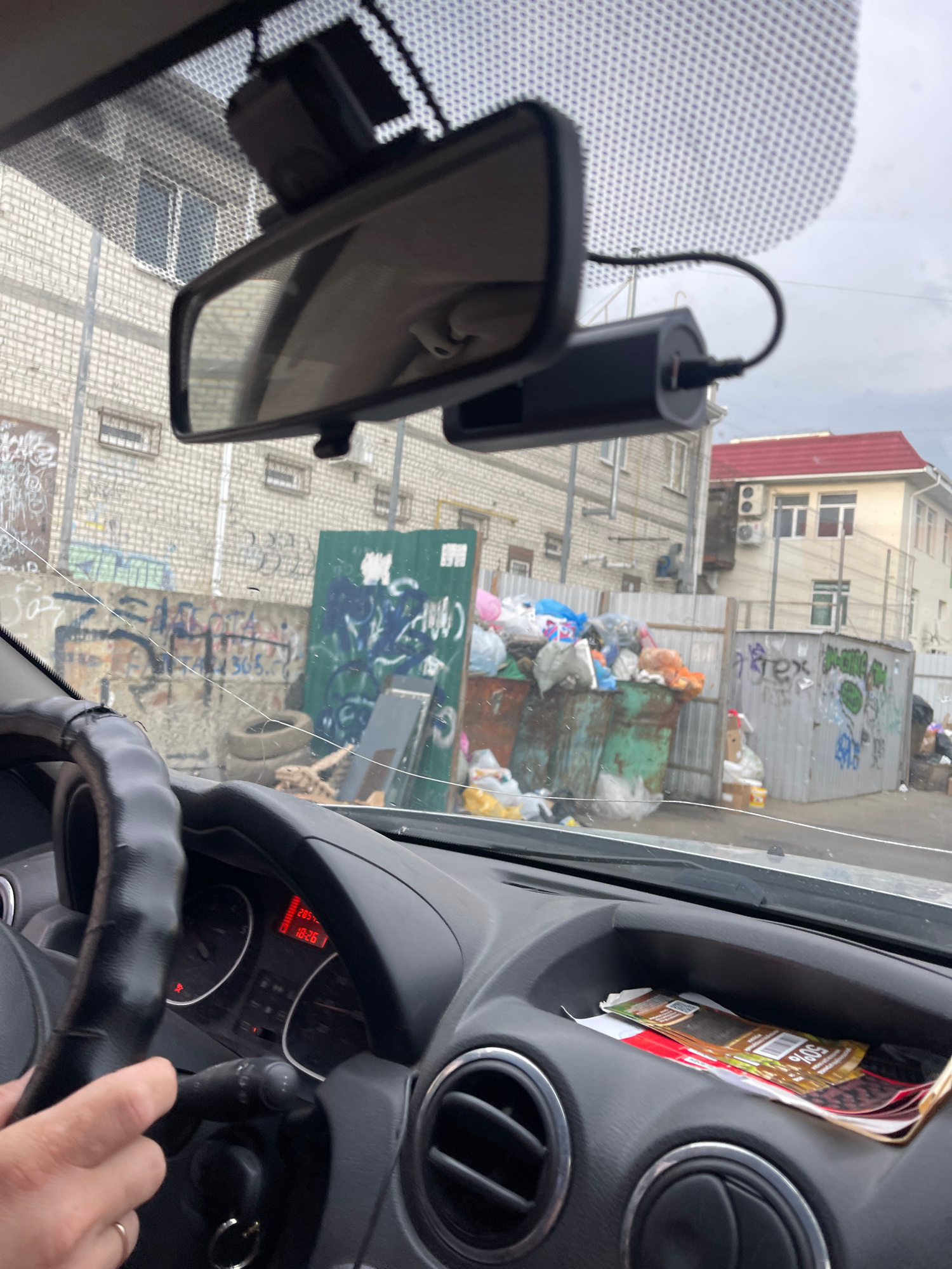 The image, captured from inside a vehicle, offers a view through the windshield. In the foreground, the steering wheel and dashboard are visible, with a hand resting on the wheel. A dashcam is mounted near the rearview mirror. Outside the vehicle, the scene depicts an urban environment characterized by dilapidated buildings, graffiti-covered walls, and overflowing dumpsters filled with garbage. The presence of a cracked windshield suggests potential damage or wear. The time displayed on the car's dashboard indicates 18:26. The context suggests a possibly neglected or run-down area within the city of Stavropol, Russia.