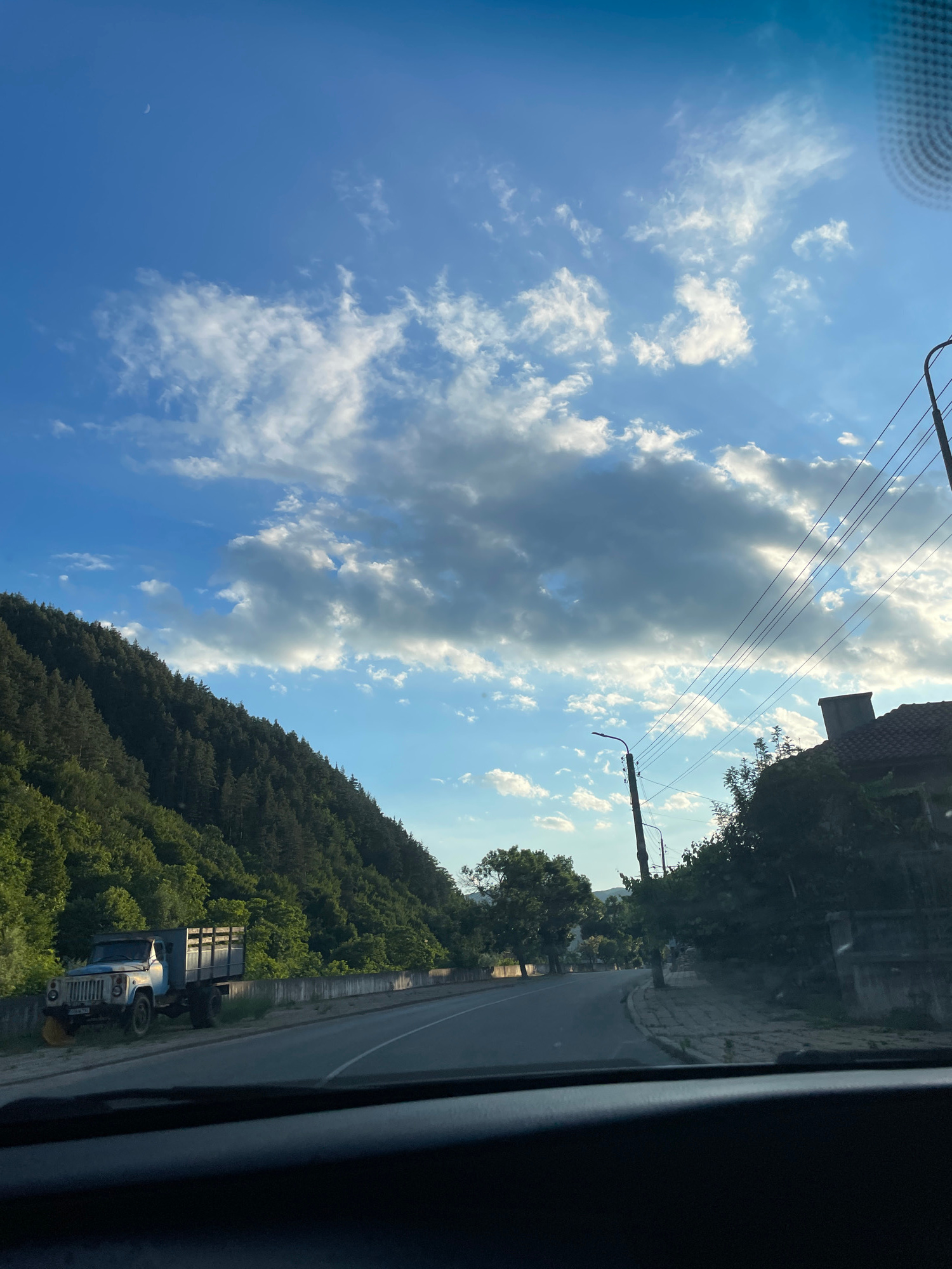An external, medium shot captures a daytime scene from a vehicle's perspective, looking forward and slightly left. The composition is dominated by a crescent moon visible in the bright blue sky, adorned with scattered white clouds. On the left, a densely wooded mountain slope slopes downwards. A light blue truck with a wooden bed is parked on the side of a paved road that curves into the distance. A concrete barrier runs parallel to the road, separating it from the truck and the mountain. On the right side of the road, a sidewalk is visible, with a house featuring a tiled roof and a chimney. Telephone poles and wires are strung across the upper right portion of the image. The overall environment suggests a rural or semi-urban setting in Zlatograd, Bulgaria.
