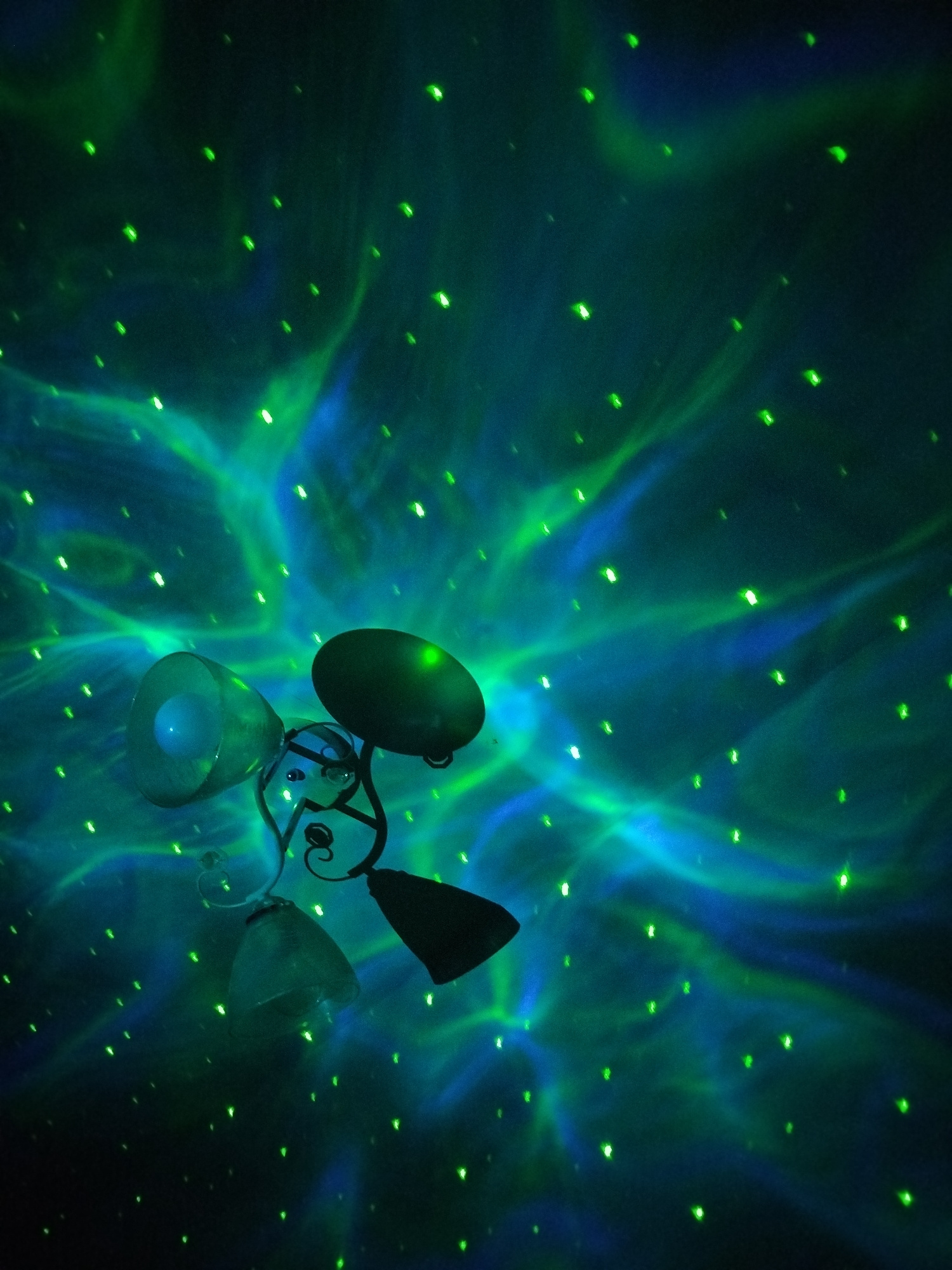 The image depicts a ceiling illuminated by a projector creating a nebula-like effect with green and blue swirls and numerous small green dots resembling stars. In the lower left quadrant, a black metal lamp fixture with three shades is visible. One shade is translucent and appears to have a light source within. The other two shades are dark and opaque. The lamp has decorative scrolled metalwork. The overall impression is of a dimly lit room with a projected celestial or cosmic light show. The location is identified as Kryvaselski Sel'ski Sovet, Belarus. There are no people or discernible actions depicted.