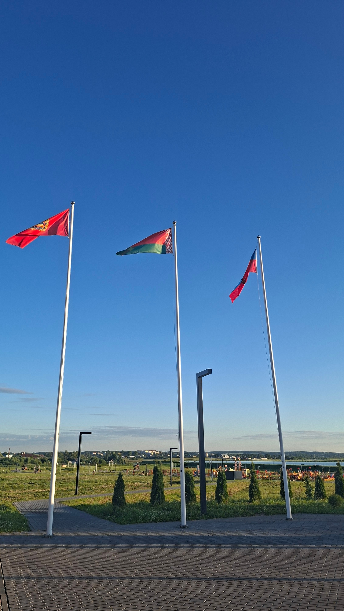 The image depicts a clear, bright blue sky with three flags flying from tall poles. The flags appear to be national or regional flags, with one clearly showing the red and green pattern of the flag of Belarus. Another flag is primarily red with a crest or emblem. The third flag is red and appears to have a blue section.
The flags are positioned at varying heights and angles, suggesting a slight breeze. Below the flags, the scene transitions to a paved walkway made of dark gray brickwork. Beyond the walkway, there is a green field with scattered trees and some playground equipment or park structures. In the distance, a body of water can be seen, possibly a lake or river, and beyond that, a few buildings are visible. The time of day appears to be late afternoon or early evening, given the long shadows cast by the structures in the field. The weather is sunny and clear. The location is identified as Smolevichi, Belarus. There is no visible text in the image. The overall impression is one of a pleasant, open-air public space on a clear day.