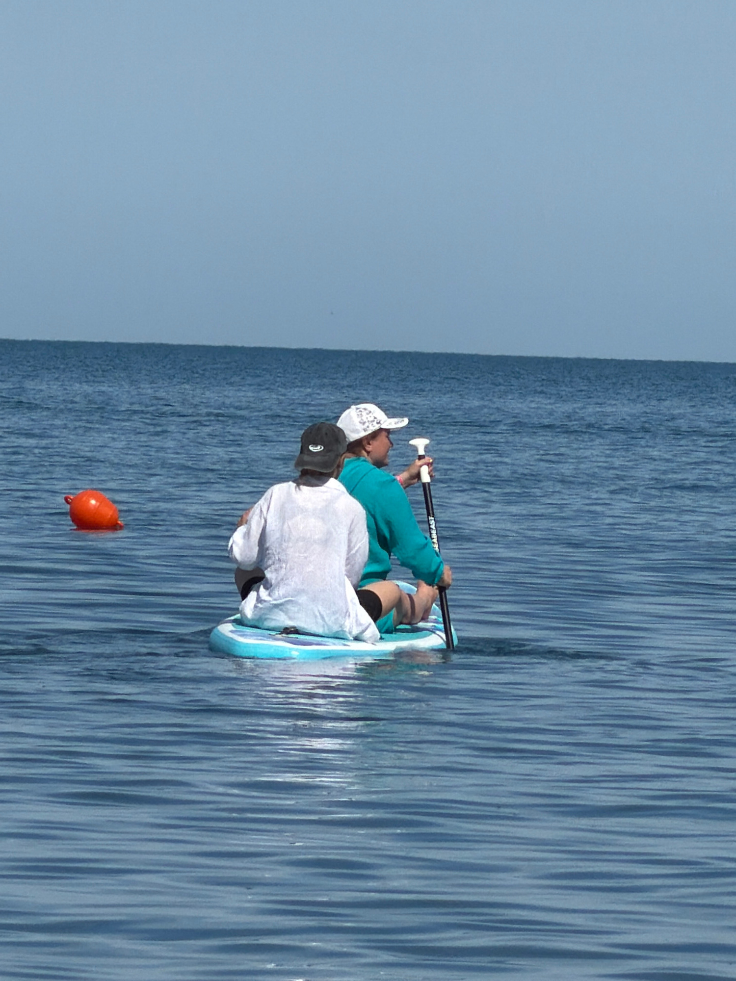 In this horizontally oriented, vertically shot image, two individuals are depicted on a paddleboard in the water. The person in the foreground, wearing a white t-shirt and a black baseball cap, is seated on the paddleboard. Their back is to the camera, and they are looking towards the left. In the background, another person is also seated on the paddleboard, wearing a teal shirt and a white patterned baseball cap. They are holding a paddle, with the handle in their right hand.
The paddleboard is light blue with white accents. The water is a deep blue with noticeable ripples. To the left of the paddleboard, a bright orange buoy floats on the surface of the water. The sky is a pale, overcast blue. The scene appears to be set during the daytime, with diffused light suggesting an overcast or early morning/late afternoon time. There is no clear indication of the specific location beyond it being a body of water. The people's postures suggest they are either resting or preparing to paddle. Visible text on the paddle reads "SEABEAST".