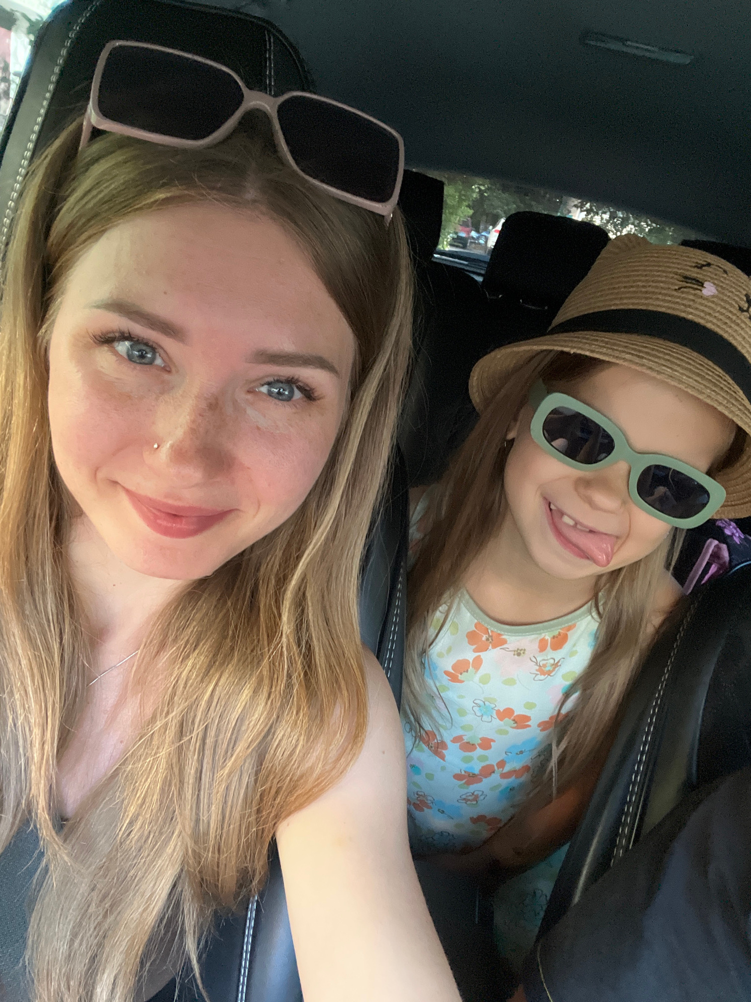 A selfie taken from inside a car shows two individuals, an adult female and a young child, likely mother and daughter, looking at the camera. The adult is in the foreground, with her blonde hair styled down and sunglasses perched on her head. She has blue eyes and freckles, and is smiling. The child is in the background, wearing a straw hat and light green sunglasses. She has long blonde hair and is sticking her tongue out, with a playful expression. She is wearing a light blue sleeveless top with an orange and floral print. The car interior is visible, with dark seats and stitching. Outside the car windows, trees and foliage are visible, suggesting an outdoor location. The presence of trees and foliage is consistent with many areas in Stepnogorsk, Kazakhstan, which has a mix of urban and green spaces.