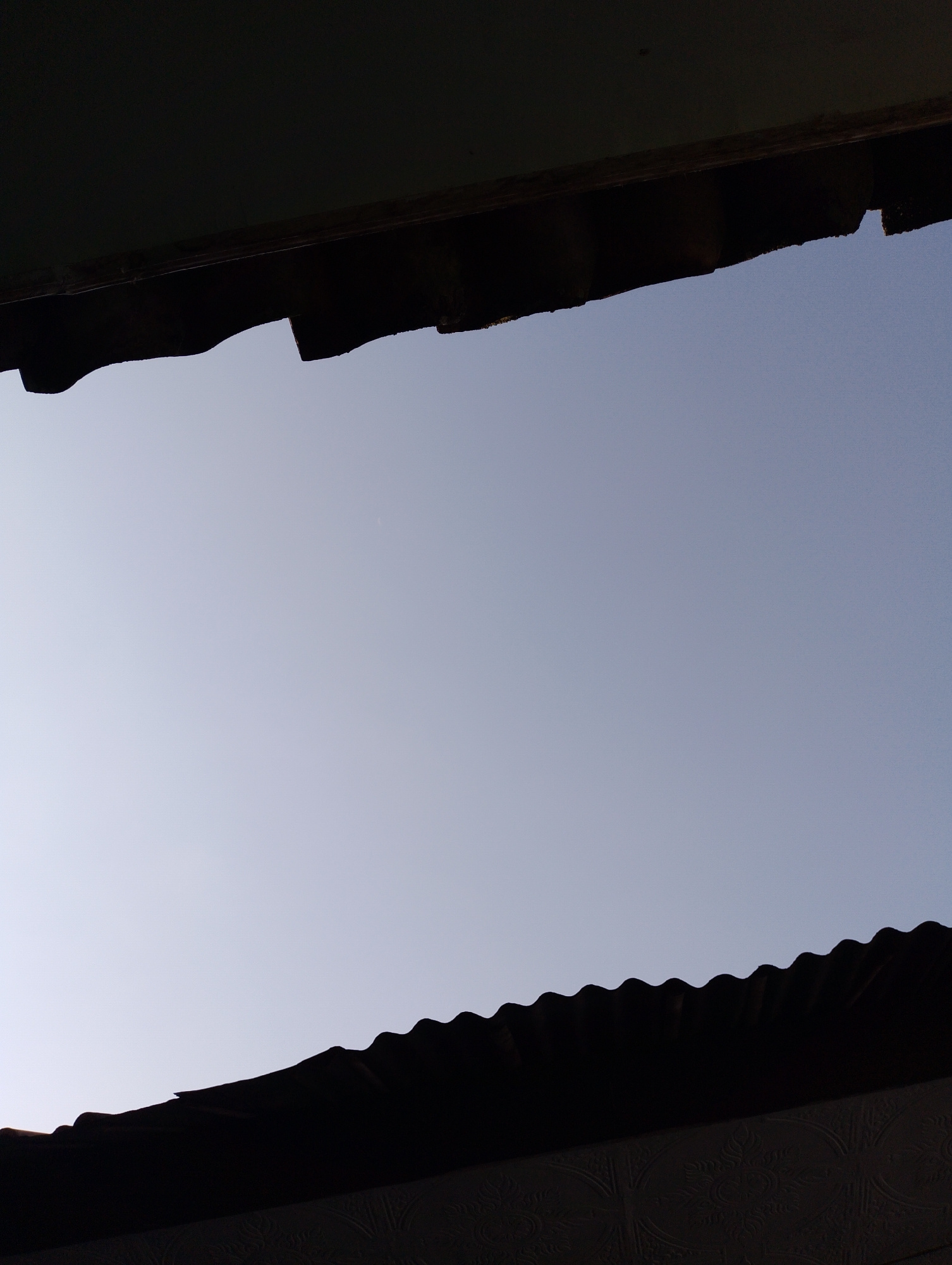 This is a low-angle, full shot of a clear sky with two dark, corrugated structures framing the view from the top and bottom. The structures appear to be roofs or overhangs, possibly made of corrugated metal, with irregular, jagged edges. The sky is a gradient of light blue, lighter towards the horizon and slightly darker towards the top. In the upper right quadrant of the sky, a very faint, small, crescent-shaped object is visible, possibly a distant moon or a very thin cloud. The bottom structure has a more defined, wavy edge and a patterned surface below it, suggesting it is part of a building. The overall scene evokes a sense of looking up from beneath an elevated structure. The location is Sukamanah, Indonesia.