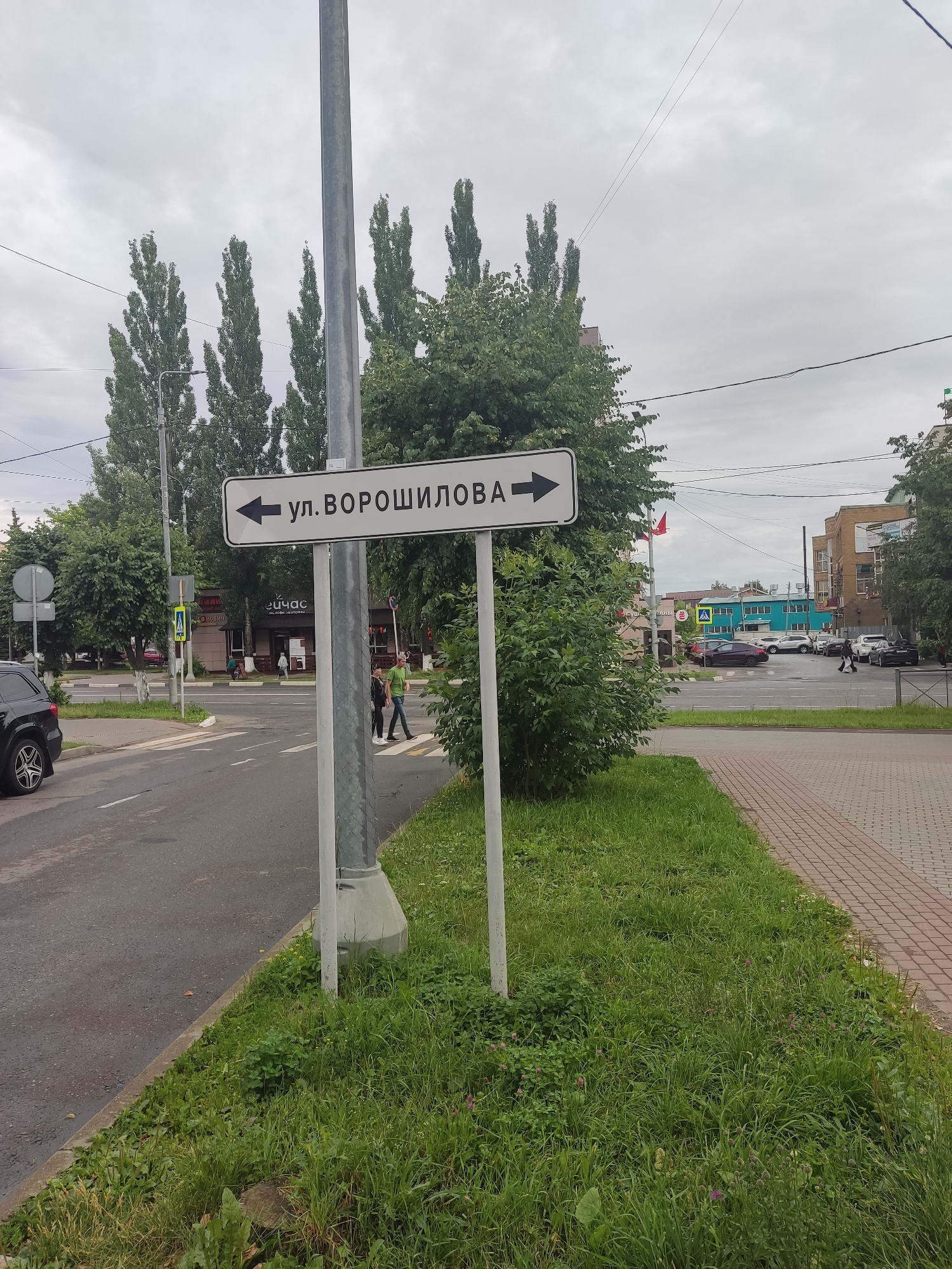 The image depicts a street scene in Serpukhov, Russia, featuring a directional signpost. The sign, oriented horizontally, indicates "ул. ВОРОШИЛОВА" with arrows pointing left and right, suggesting two directions for this street. The signpost is a metal pole with a concrete base, positioned on a patch of grass next to a paved road.
In the background, there are several tall trees with lush green foliage, suggesting a temperate climate. Buildings of various architectural styles are visible, including a light brown, multi-story building and a lower, bright turquoise building. A street with several cars parked and in motion is also present.
People are visible on a crosswalk in the mid-ground, crossing the street. One group consists of two individuals walking together, and another person is crossing further down. A black car is parked on the left side of the road, partially visible in the foreground. The sky is overcast, with a cloudy gray sky. The scene appears to be a typical urban environment on a cloudy day.