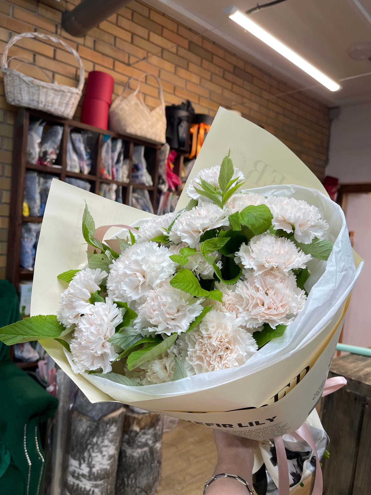 A person's hand holds a bouquet of white carnations. The bouquet is wrapped in light yellow and white paper with pink ribbon accents. Green leaves are interspersed among the flowers. The background is out of focus but appears to be a room with shelves filled with various items, including baskets and rolls of fabric. The wall behind the shelves is made of brick. A bright fluorescent light fixture is visible on the right side of the image. The scene is indoors and appears to be in a retail or storage space. There is some text visible on the wrapping paper, but it is partially obscured and difficult to read clearly. The overall impression is a candid shot of a person holding a floral arrangement.