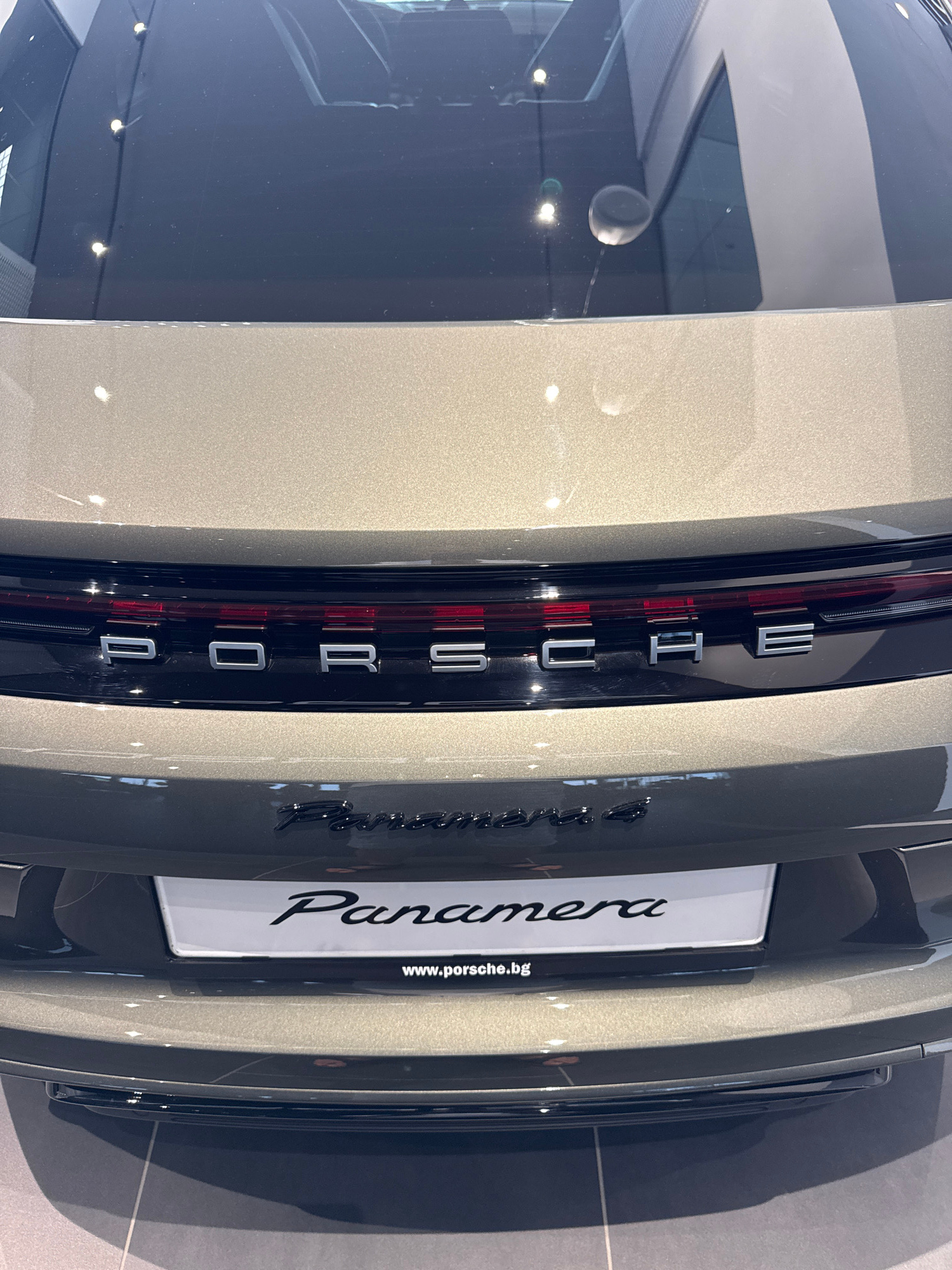 An eye-level, medium shot focuses on the rear of a metallic bronze Porsche Panamera. The car's "PORSCHE" emblem is prominently displayed in silver chrome across the dark taillight bar. Below, the "Panamera" model name is written in a stylized script on a white license plate, with the website "www.porsche.bg" visible at the bottom. The background shows a showroom interior, indicated by the polished floor tiles and subtle overhead lighting reflected on the car's surface. The setting is the Porsche Center Sofia, located in Radinovo, Bulgaria. There are no people or animals present.