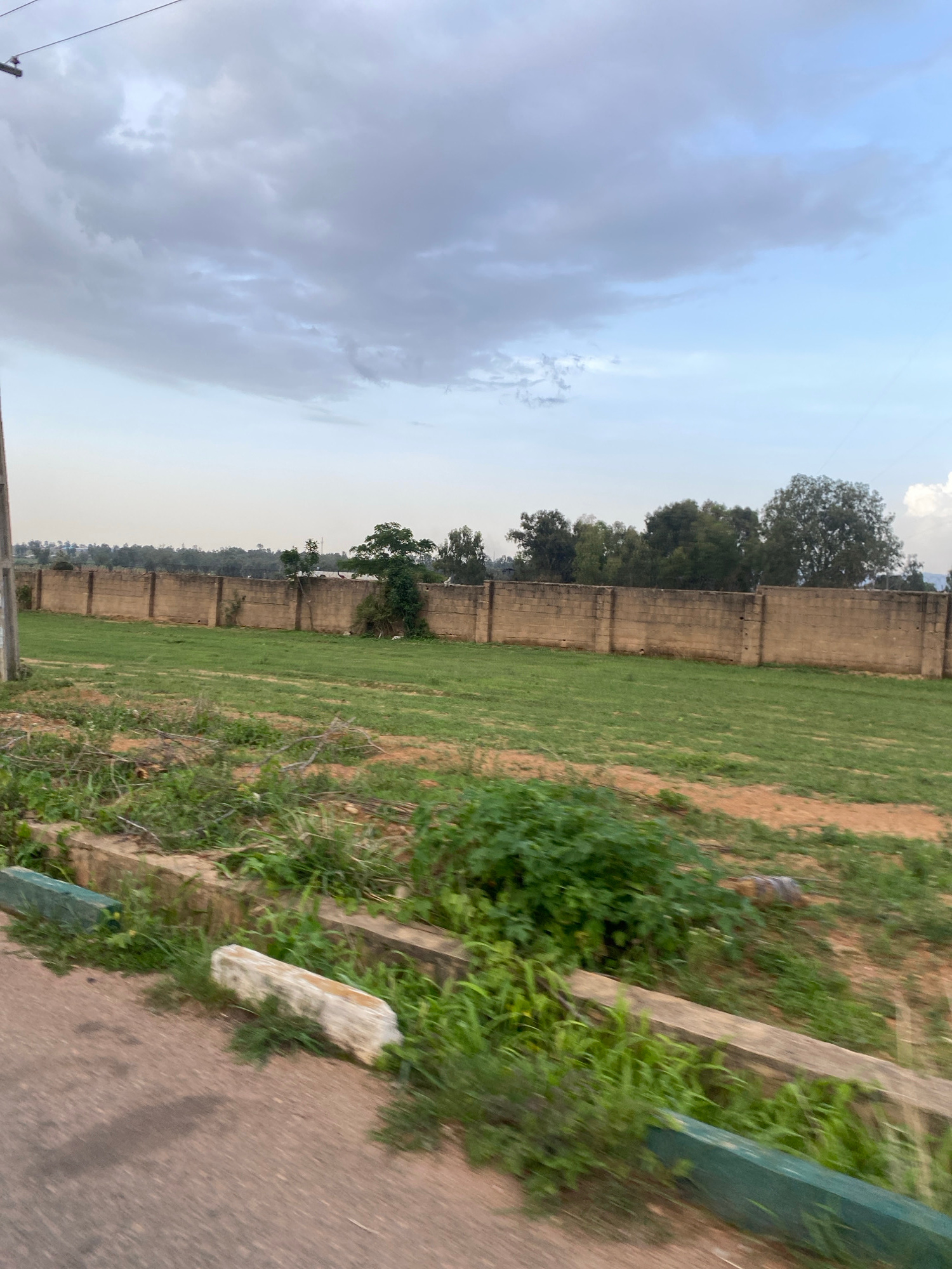 A low-angle, slightly blurred, daylight, medium shot captures a long, tan, cinder block wall stretching horizontally across the mid-ground of the frame, dividing a green, grassy field from a distant tree line. The wall is punctuated by vertical pillars and shows signs of weathering. A power pole is visible on the far left. The field in front of the wall is a mix of green grass and bare earth, with some green foliage growing in clumps and along the edge of a road in the foreground. The road surface is a reddish-brown asphalt. The sky above is overcast with dramatic grey and white clouds. The location is the outskirts of Barikin Jirga Summa, Nigeria.