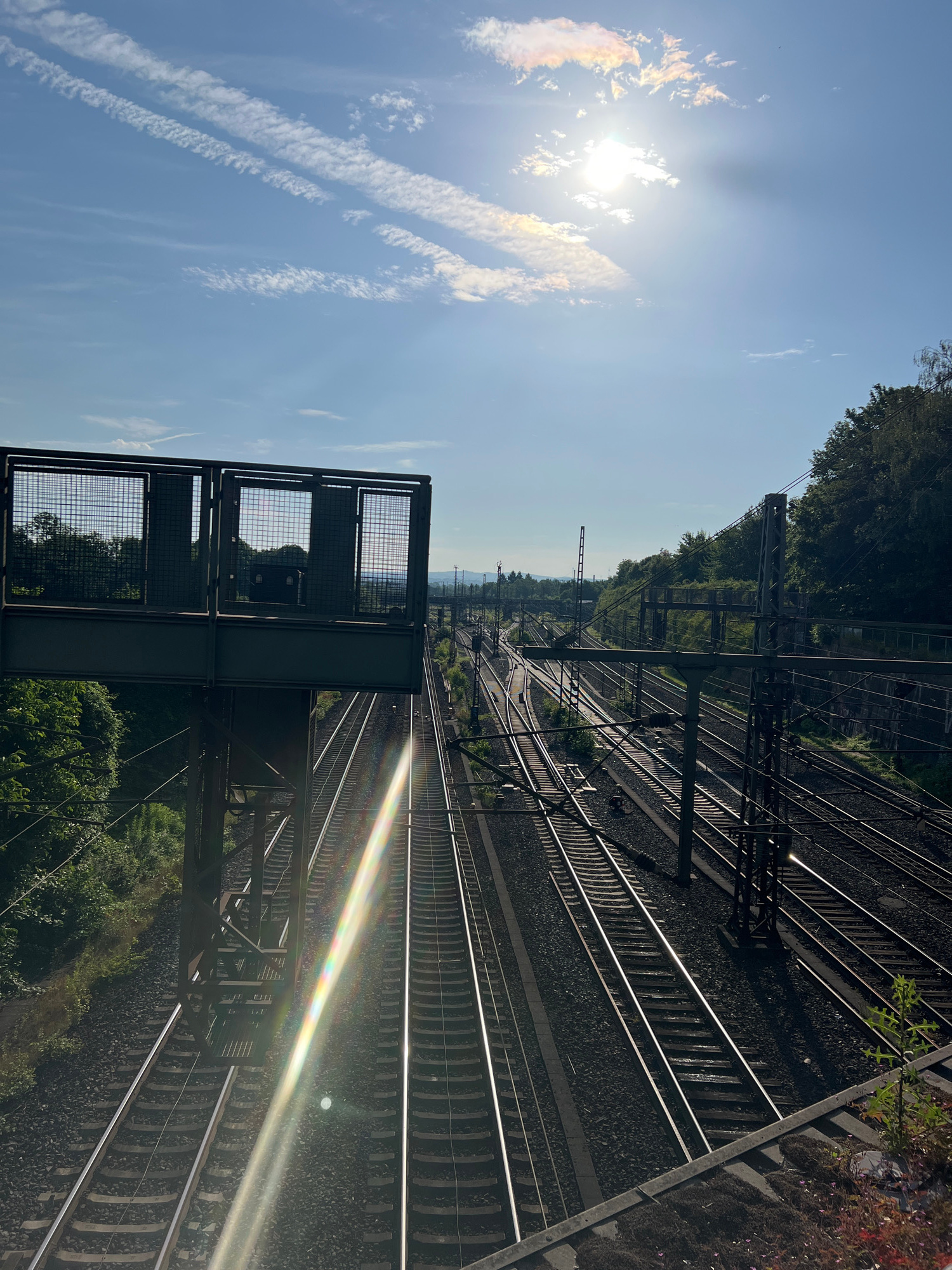 A sun-drenched, slightly hazy sky with wispy clouds hangs over a complex railway junction. In the foreground, a metal pedestrian overpass with a wire mesh railing extends over multiple sets of train tracks. The tracks diverge and converge, disappearing into the distance. Sunlight glints off the rails and creates a strong lens flare, rainbow effect, in the lower left portion of the image. Lush green trees line the sides of the tracks, and in the far distance, rolling hills are visible under the bright sun. The scene is devoid of people, and the focus is on the infrastructure of the railway and the natural elements.