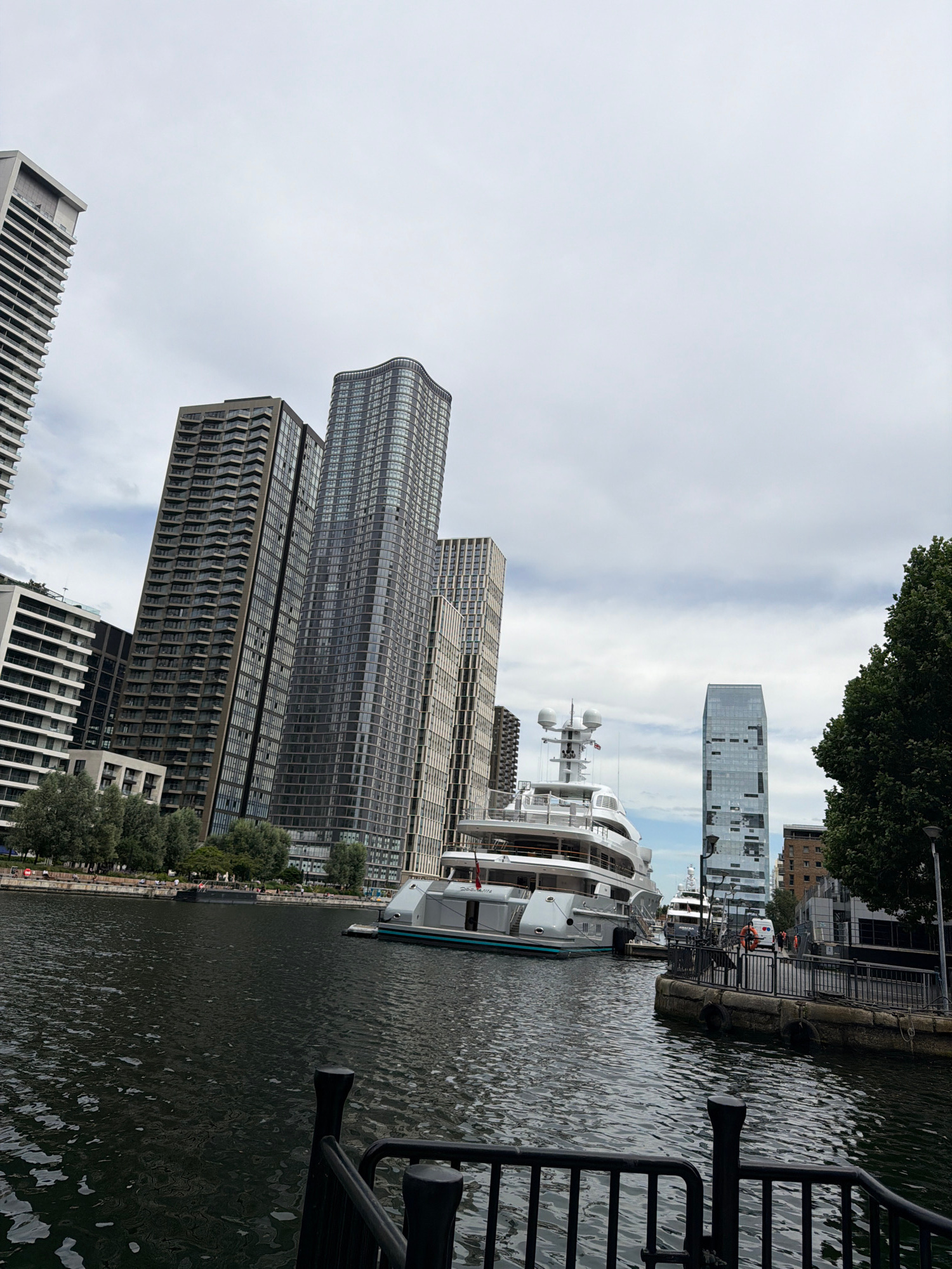 The image depicts a waterfront scene in London, United Kingdom. In the foreground, a dark body of water is visible, with a black railing in the lower portion of the frame. Several large, modern buildings with glass facades dominate the background, varying in height and architectural style. A large white yacht is moored in the water, and other smaller vessels are also present. The sky is overcast with clouds. People can be seen walking along the waterfront promenade. The overall impression is one of a contemporary urban environment with a focus on the river and modern architecture.