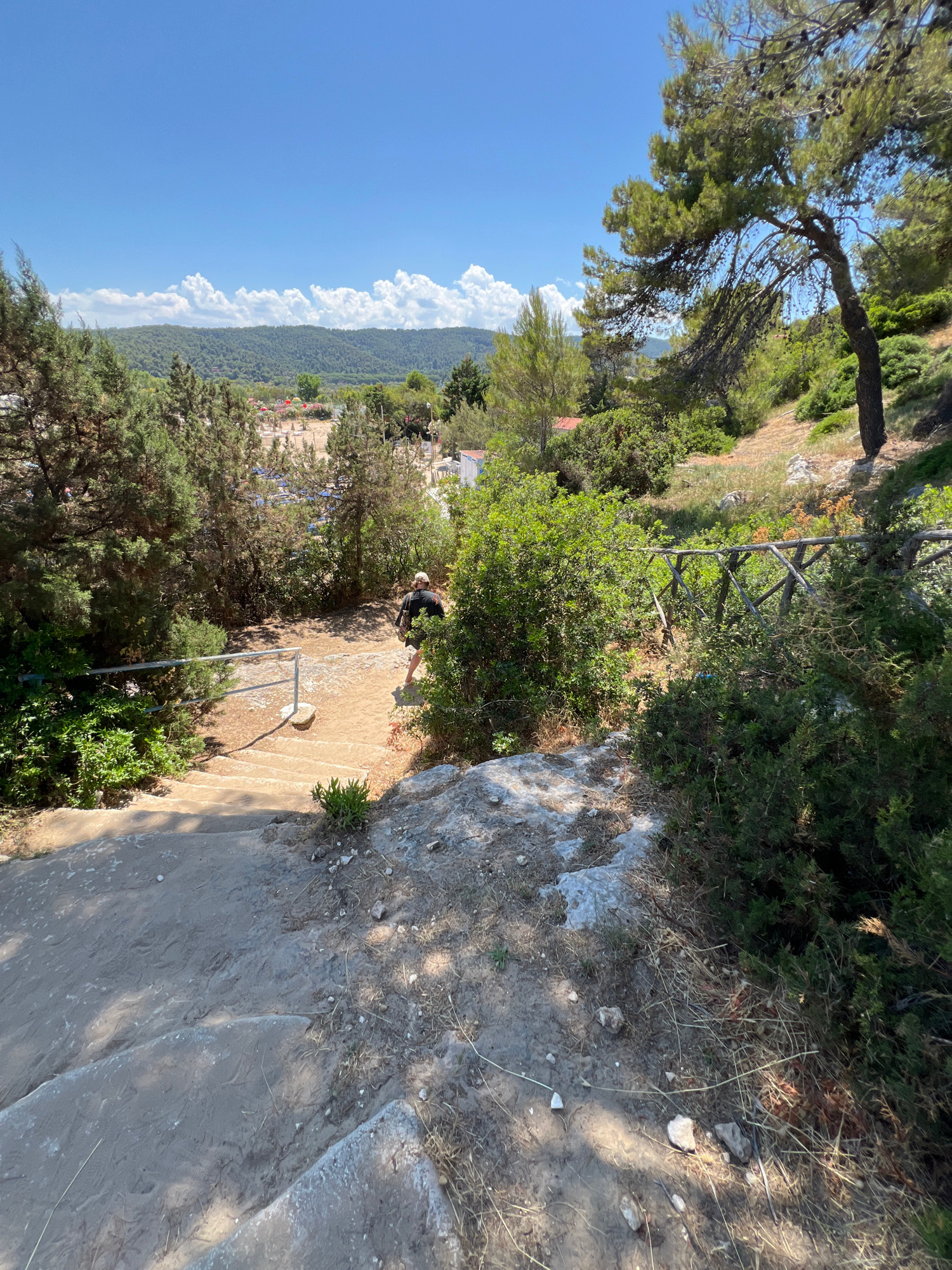 The image shows a scenic view of a path leading down towards a beach in Peschici, Italy. The path is made of concrete steps and sandy terrain, bordered by lush green vegetation, including pine trees and bushes. A metal handrail runs along the side of the path for support. In the mid-ground, a person wearing a dark shirt and shorts is walking down the path. Beyond the path, a beach with numerous rows of lounge chairs and umbrellas is visible, situated near a body of water, likely the Adriatic Sea. In the background, rolling green hills covered in trees form a natural backdrop under a clear blue sky dotted with white clouds. The overall scene depicts a picturesque and inviting coastal environment, characteristic of the Peschici region.