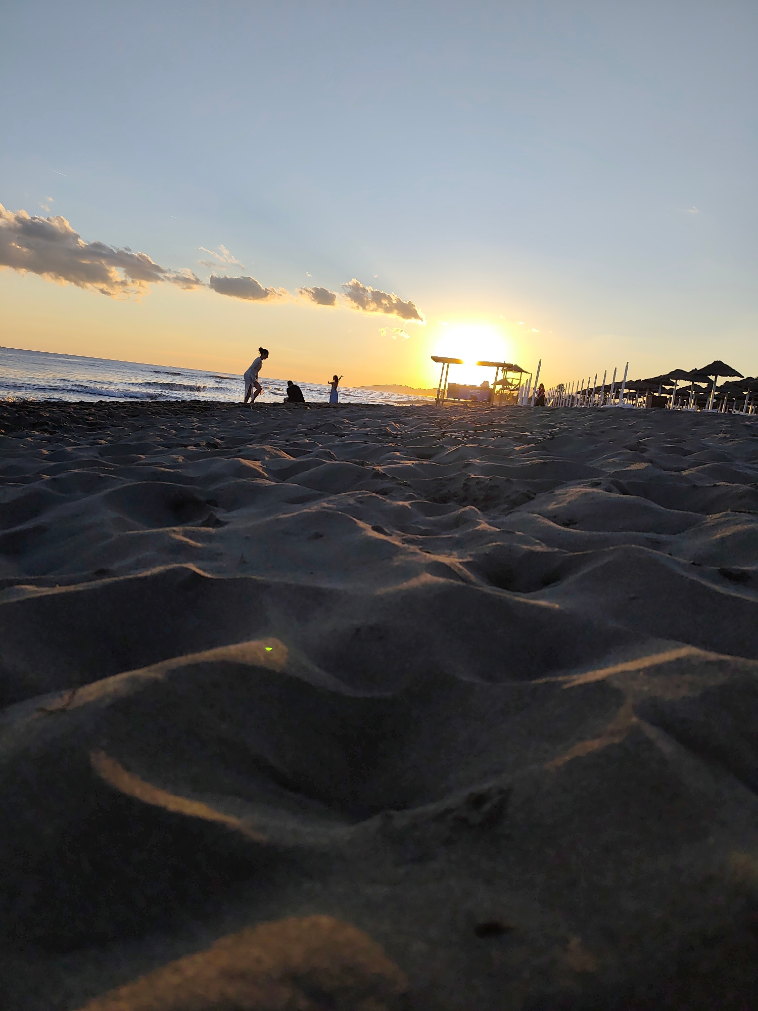 The image captures a sunset scene on a sandy beach in Donji Štoj - Shtoji i Poshtëm, Montenegro. In the foreground, the sand is textured with dunes and shadows, with a small green reflection visible on a sand dune. In the mid-ground, several people are present on the beach. A woman in light-colored clothing is standing and facing left, possibly playing with or observing children. Two children are also visible; one is closer to the woman and appears to be playing, while the other is further away and appears to be running or jumping. In the background, the sun is setting on the horizon, casting a warm glow. There is a line of beach umbrellas and lounge chairs, with some structures that may be beach huts or lifeguard stations. The sea is visible behind the people, with gentle waves. The sky is a gradient of blue and orange, with scattered clouds.