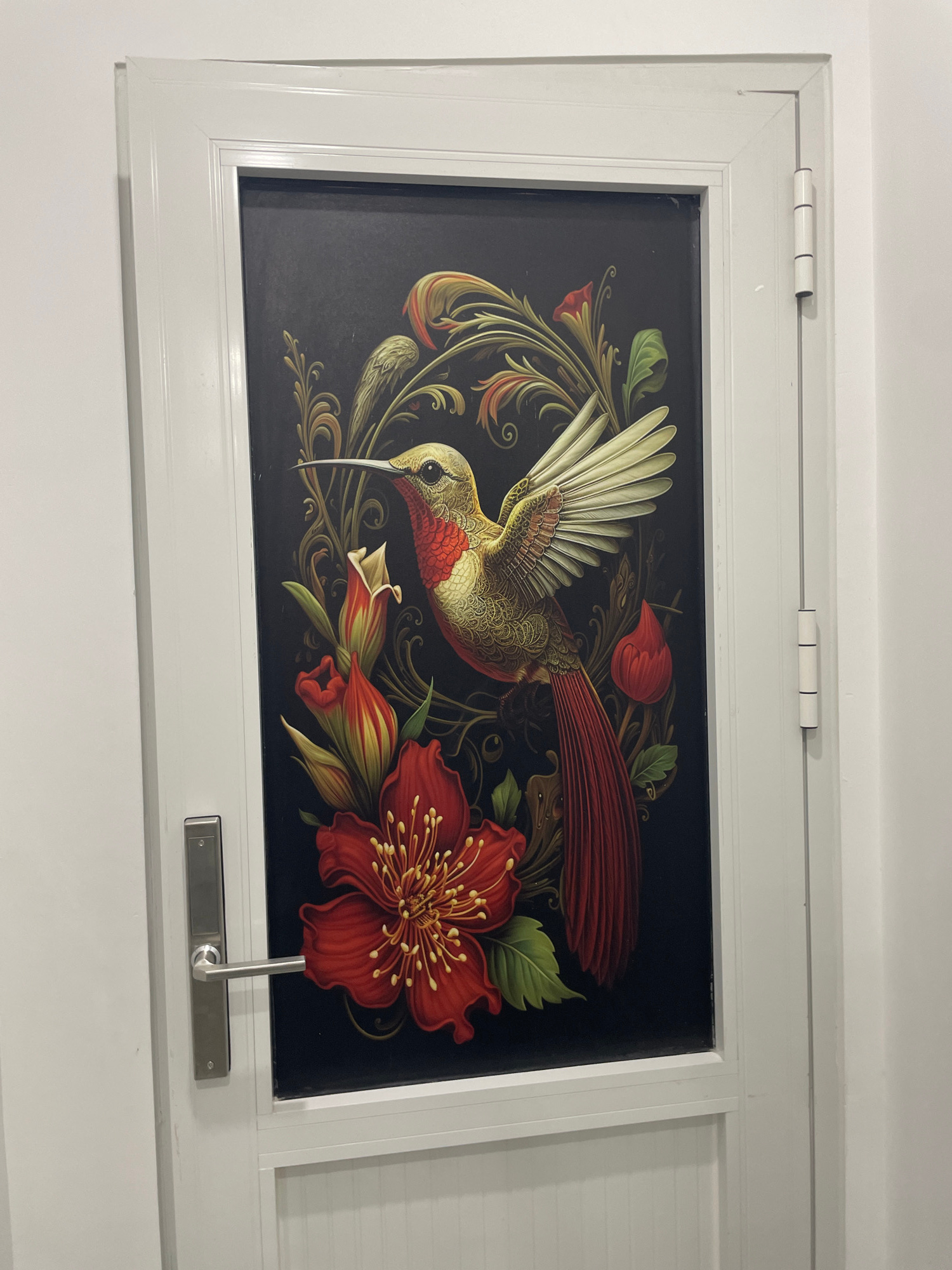 The image shows a white door with a decorative panel featuring a stylized depiction of a hummingbird in flight amidst red flowers and ornate foliage against a black background. The door has a silver handle and is hinged on the right side. The scene is set in Thuận An, Vietnam, as indicated by the metadata. The environment appears to be indoors, possibly a residential or commercial space, given the clean, well-lit appearance of the door and surrounding wall. There are no people visible in the image.