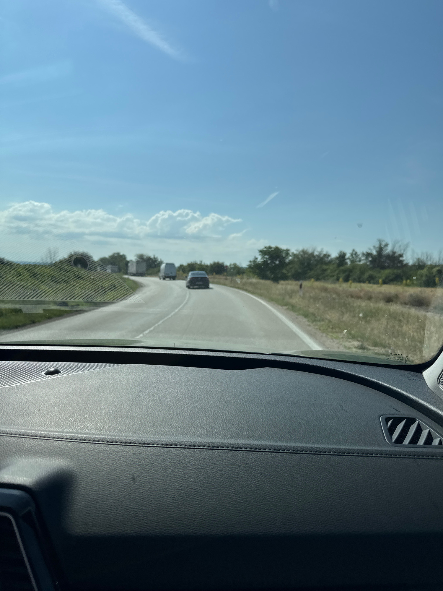 The image captures a view from inside a vehicle looking forward through the windshield. The road ahead is asphalt and curves gently to the right. There are several vehicles on the road, including a dark-colored car in the immediate foreground, followed by a white van, and further ahead, two white trucks. The landscape on either side of the road is rural, with dry, grassy fields and scattered trees. The sky is a bright blue with some scattered white clouds. The foreground of the image shows the dashboard of the vehicle, which is dark gray and textured, with stitching detail. This scene is consistent with a highway or rural road in the city of Archar, Bulgaria.