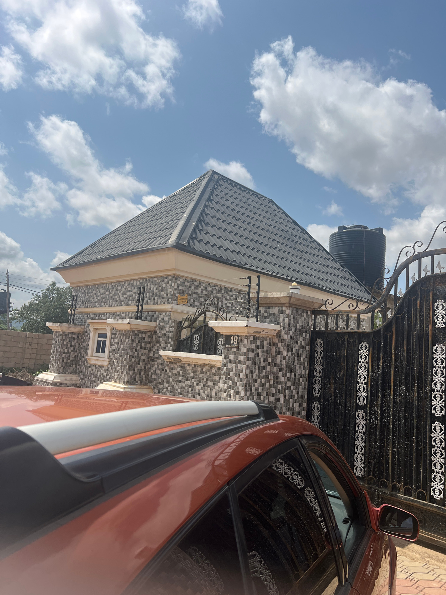 The image shows a close-up, Dutch angle view of a building's facade and a portion of an orange car. The building features a dark, corrugated metal roof and walls clad in small, rectangular mosaic tiles in shades of grey and white. A black water tank is visible on the roof, and an ornate black metal gate with scrollwork is in the background. A small white-framed window is set into the tiled wall. The car's side, including its side mirror, is prominent in the foreground, reflecting the sky and the building.
The sky is blue with scattered white, puffy clouds, suggesting it is daytime. The lighting indicates a bright, sunny day. The image appears to be taken outdoors, in what seems to be a residential or commercial area, given the presence of a house and a gate. The scene is static, with no discernible activity or event taking place.
A number "18" is visible on a white rectangular plaque mounted on the building wall. The overall impression is that of a brightly lit, possibly warm, day in a built-up area in Howayi, Nigeria.