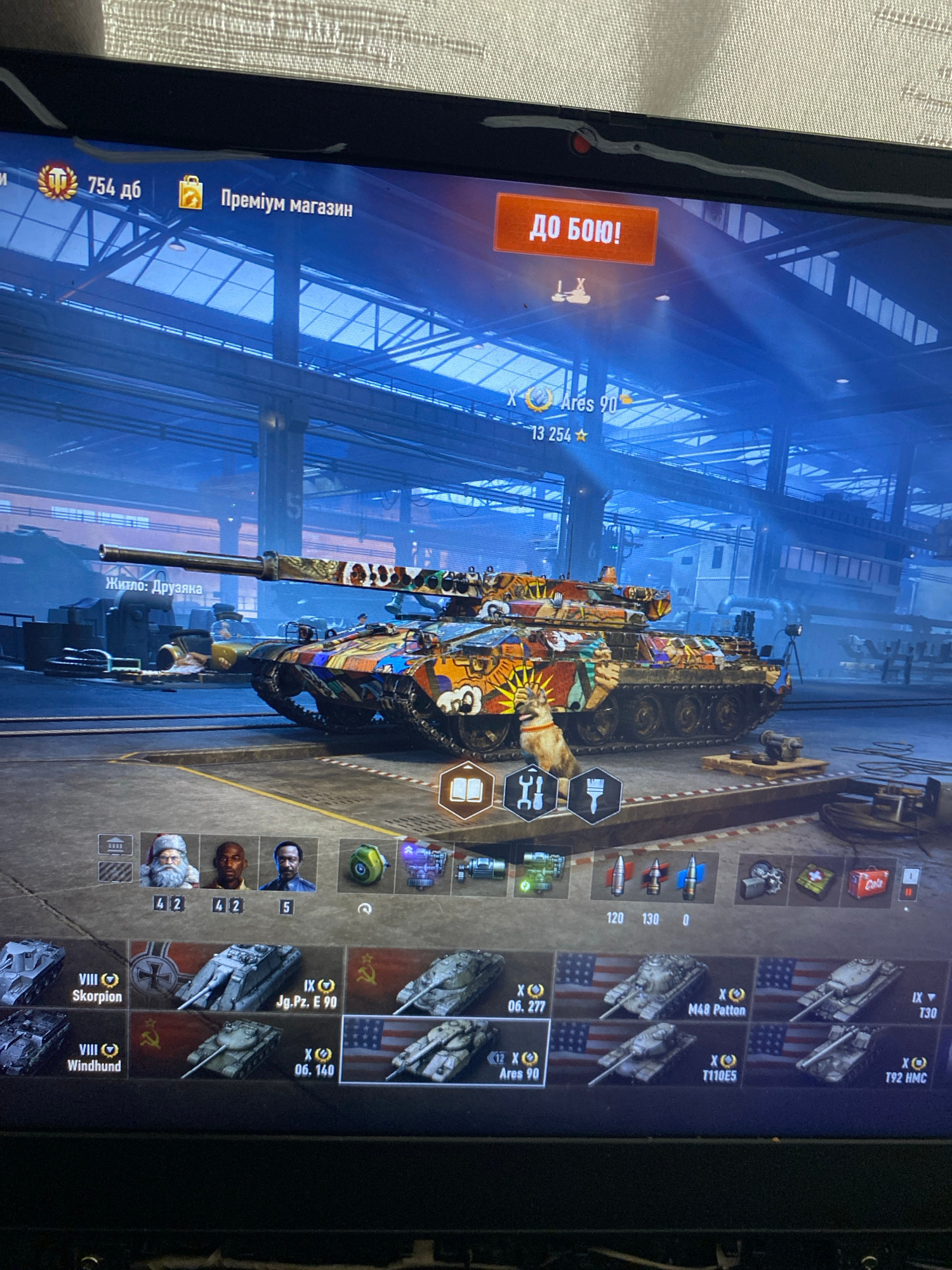 The image shows a computer screen displaying a video game interface, likely a tank combat game. The game is in Russian, as indicated by the text "ПРЕМІУМ МАГАЗИН" (Premium Shop) and "ДО БОЮ!" (To Battle!). The main subject is a heavily customized tank with a vibrant, cartoonish paint job, parked in what appears to be a virtual garage or hangar environment. The interface shows various game-related icons and information, including vehicle tiers, names, and player avatars. There are no discernible features or elements in the image that would allow for the identification of a specific city or location, including Szczecin, Poland. The scene is entirely digital and contained within the computer display.