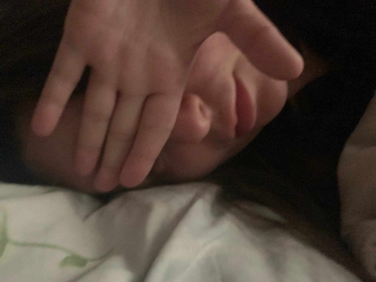 A close-up, low-light, top-down view shows a person's face partially covered by their hand. The person is lying on a white surface with green markings, possibly a sheet or duvet. The lighting suggests an indoor environment, likely at night or in a dark room. The focus is on the hand and a portion of the face, with the background blurred and dark. The image is grainy due to the low light conditions.