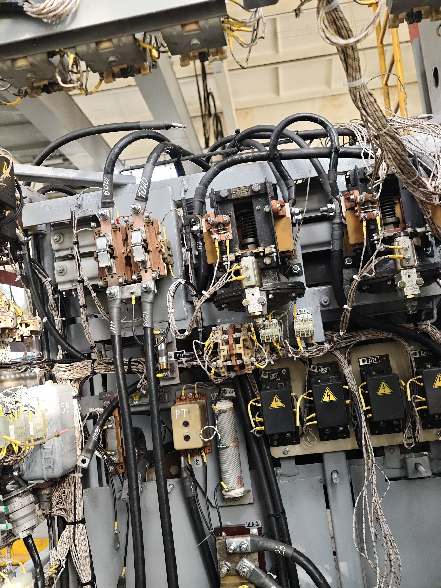This image is a close-up, low-angle shot of a complex industrial electrical panel. The panel is filled with a dense arrangement of wires, cables, circuit breakers, relays, and other electrical components. Thick black cables are prominent, some labeled with numbers like "1009", "7010", "1005", and "1008". Smaller, multicolored wires are bundled together, secured with zip ties. Several rectangular black boxes with yellow lightning bolt symbols are visible, likely fuses or circuit protection devices. Some components have Cyrillic lettering, such as "ДБ1", "ДБ2", "ДТ1", and "ДН1". A cream-colored cylindrical object with a white band and visible winding is mounted vertically, potentially a resistor or coil. The overall environment appears to be an interior industrial space, characterized by grey metal surfaces and a utilitarian aesthetic. The lighting suggests overhead industrial lighting, creating some shadows. There are no people or discernible environmental features that would indicate a specific city or location.