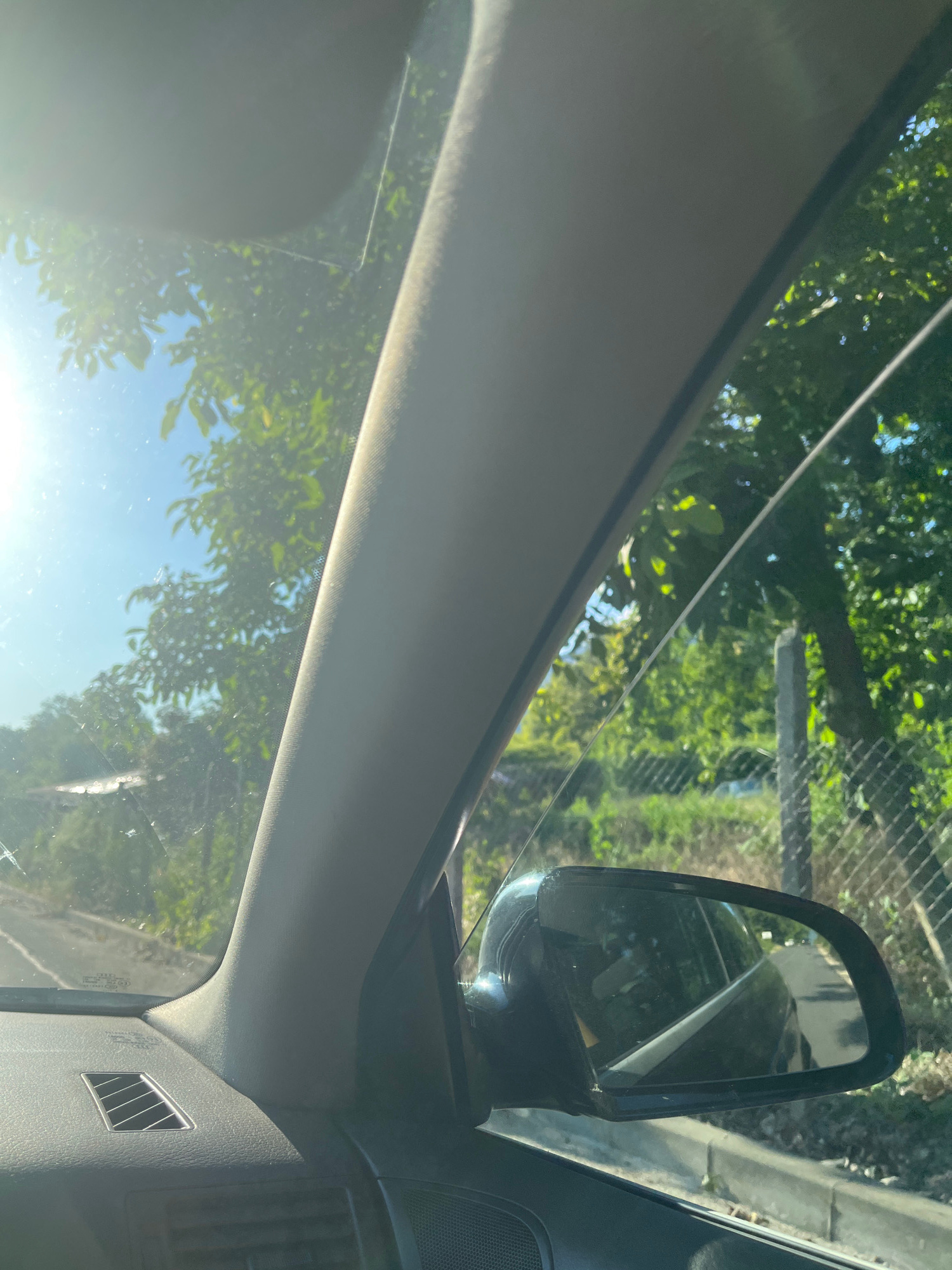 The image shows a view from inside a car, looking out the windshield and side window towards a sunny outdoor scene. The interior of the car is partially visible, including a dark gray dashboard with a vent, and a gray pillar. Outside the car, trees with green foliage and blue sky are visible. Sunlight shines brightly, creating lens flares on the windshield. A side mirror reflects the interior of the car and the road behind. A wire fence and a concrete post are also visible along the side of the road. The scene appears to be a rural or suburban road, likely during daytime.