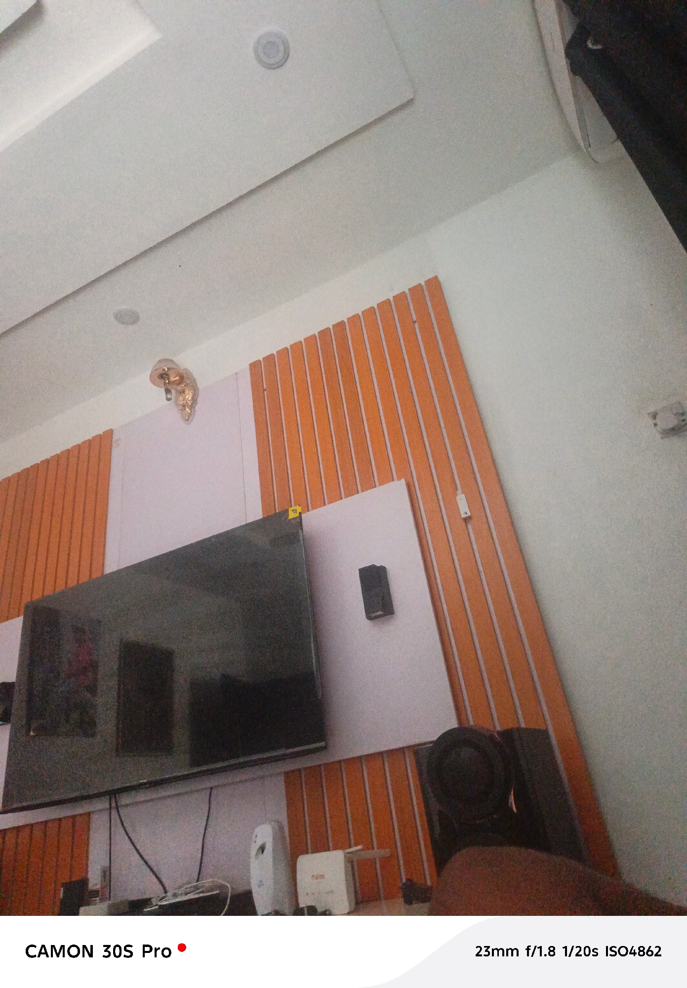 The image depicts an indoor scene, likely a living room or entertainment area. A large, dark television screen dominates the lower-middle portion of the frame, mounted on a wall that features a light purple panel bordered by vertical orange wooden slats. Above the TV, a decorative gold-colored object, possibly a sculpture or lamp, is attached to the ceiling. The ceiling itself is white with a modern, multi-level design, and contains two circular recessed lights.
To the right of the TV, a portion of a dark armchair is visible, with a person's arm and shoulder visible in the lower-right corner, suggesting they are seated in it. On the shelf below the TV, several objects are present, including a white air freshener or diffuser, a white box labeled " Airtel" which is a telecommunications company, and some wires.
The lighting suggests it is daytime, as natural light appears to be entering the room, though the primary illumination source is not clearly identifiable. The overall ambiance is casual and domestic. Text visible in the lower portion of the image includes "CAMON 30S Pro" with a red dot, and "23mm f/1.8 1/20s ISO4862", which appear to be camera settings or branding.