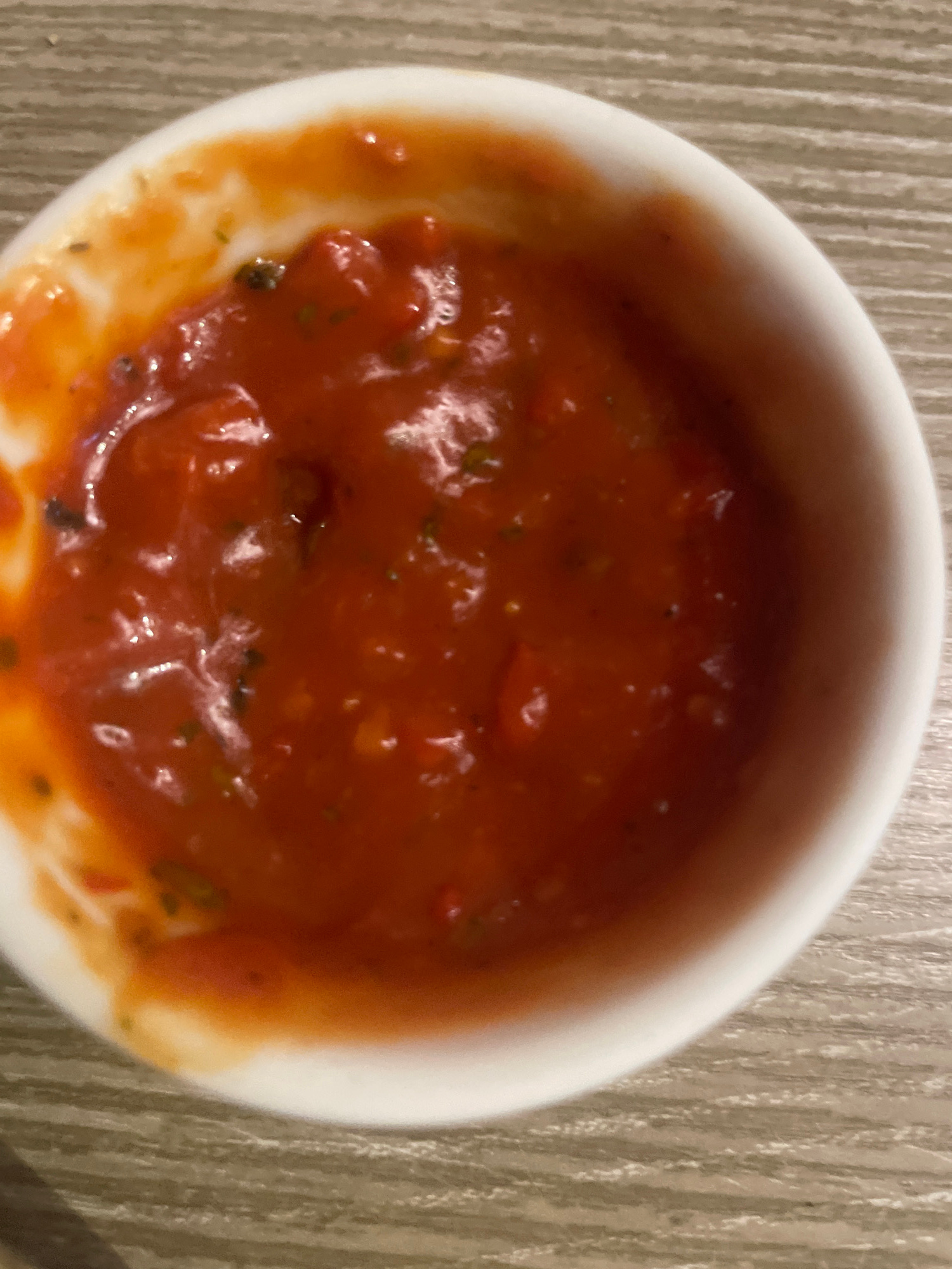 A white ceramic ramekin filled with a thick, red tomato-based sauce, likely salsa or a similar condiment, rests on a wooden table with a visible wood grain pattern. The sauce is chunky, with discernible pieces of tomato and what appear to be herbs or spices mixed throughout. The lighting is overhead, casting subtle shadows and highlighting the glossy texture of the sauce. The scene is indoors, with no indication of an outdoor setting. The city of Minsk, Belarus, is not discernible from the image's content.