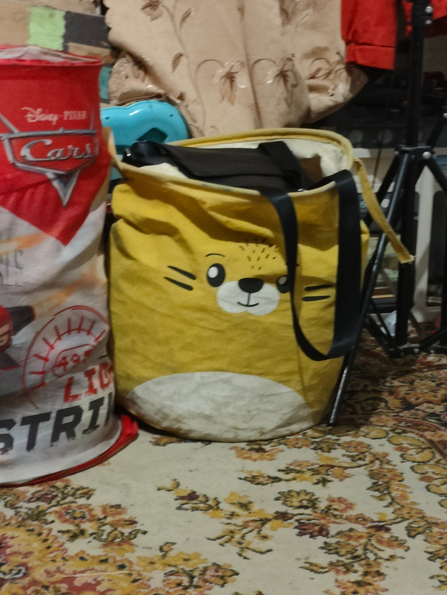 In Yarmolyntsi, Ukraine, two laundry bags stand on a patterned carpet. The left bag is red with a white design featuring the Disney Pixar "Cars" logo and the word "Cars" in silver. Below this, text in red and black is partially visible, including "49" and "STRI". The right bag is yellow and shaped like a cartoon animal's face with black eyes and a mouth, and white cheeks. It has black handles. Behind the bags, a blue plastic object and draped fabric are visible. A black metal tripod stands to the right. The overall scene suggests an indoor domestic setting.