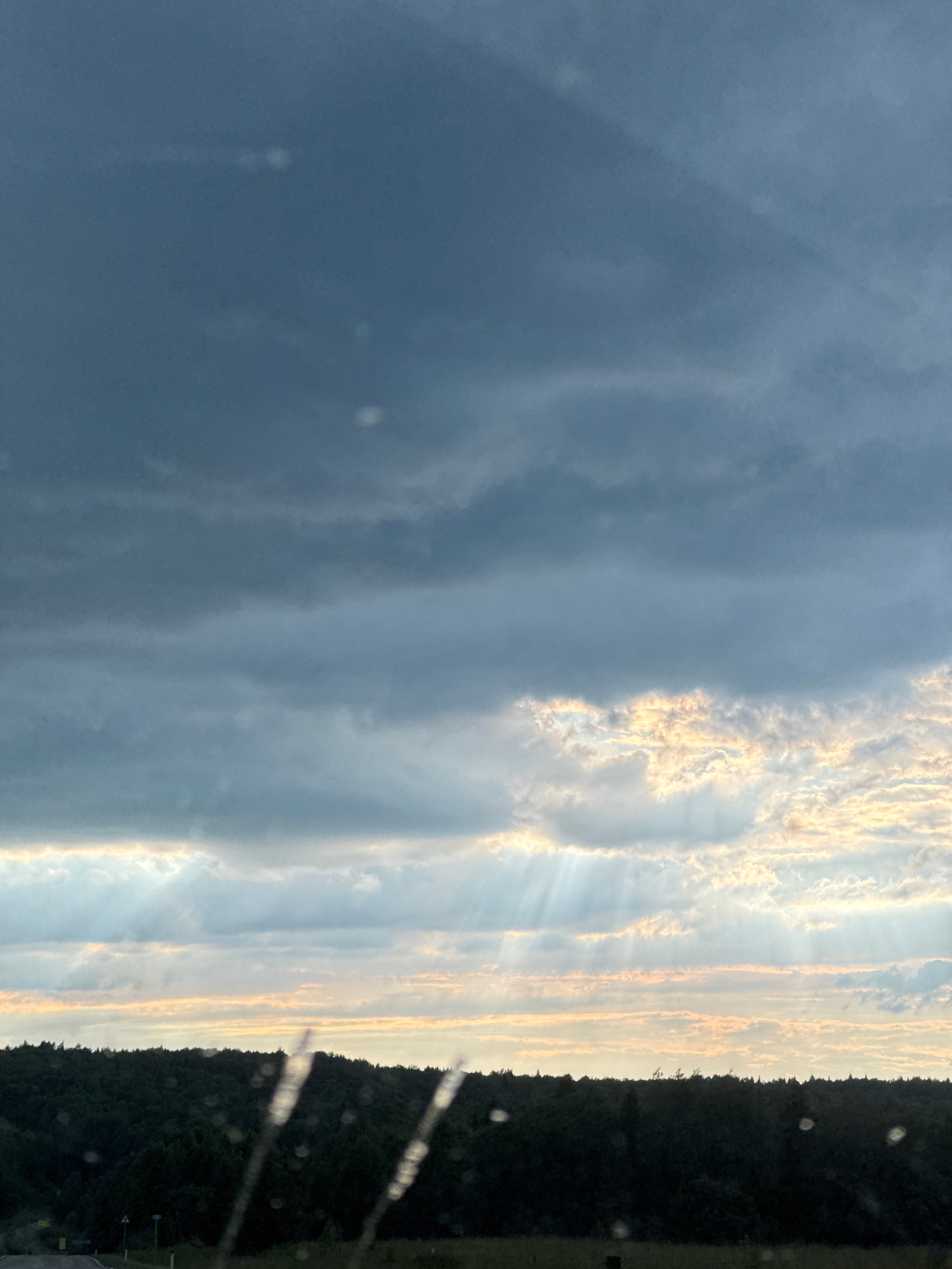 The image captures a dramatic sky over a densely forested area. Dark, heavy clouds dominate the upper portion of the frame, suggesting an approaching storm or the aftermath of one. Shafts of light, known as crepuscular rays, pierce through gaps in the clouds lower in the sky, illuminating patches of lighter, broken clouds with warm, golden hues. Below the sky, a dark, silhouetted line of trees forms a dense forest that stretches across the horizon. The foreground is blurred, with vertical streaks that appear to be water droplets or streaks on a window, hinting that the photograph may have been taken from inside a vehicle. The environment appears to be natural and rural. There are no people or specific man-made structures clearly visible in the main scene, although a faint road and signs are present in the far lower left corner. The overall mood is one of natural power and atmospheric beauty. The location is specified as Большая Уса, Russia.