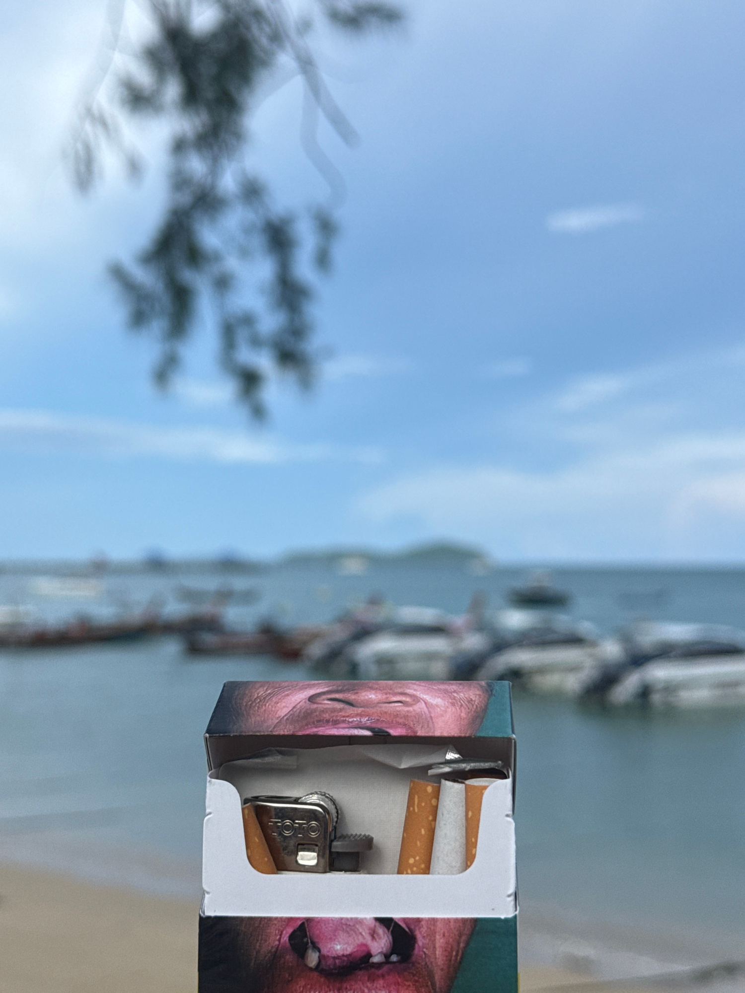 A close-up, eye-level, medium shot captures a cigarette pack with two cigarettes and a lighter inside it, positioned in the foreground. The pack is adorned with a graphic of a human face, with a mouth and eyes visible on the front and sides. The background is blurred, revealing a tranquil beach scene with boats docked on the ocean under a cloudy sky. The location is identified as Rawai, Thailand, a popular tourist destination known for its beaches and seafood restaurants. The lighting is natural, with the sun casting a soft glow on the scene, creating a peaceful atmosphere.